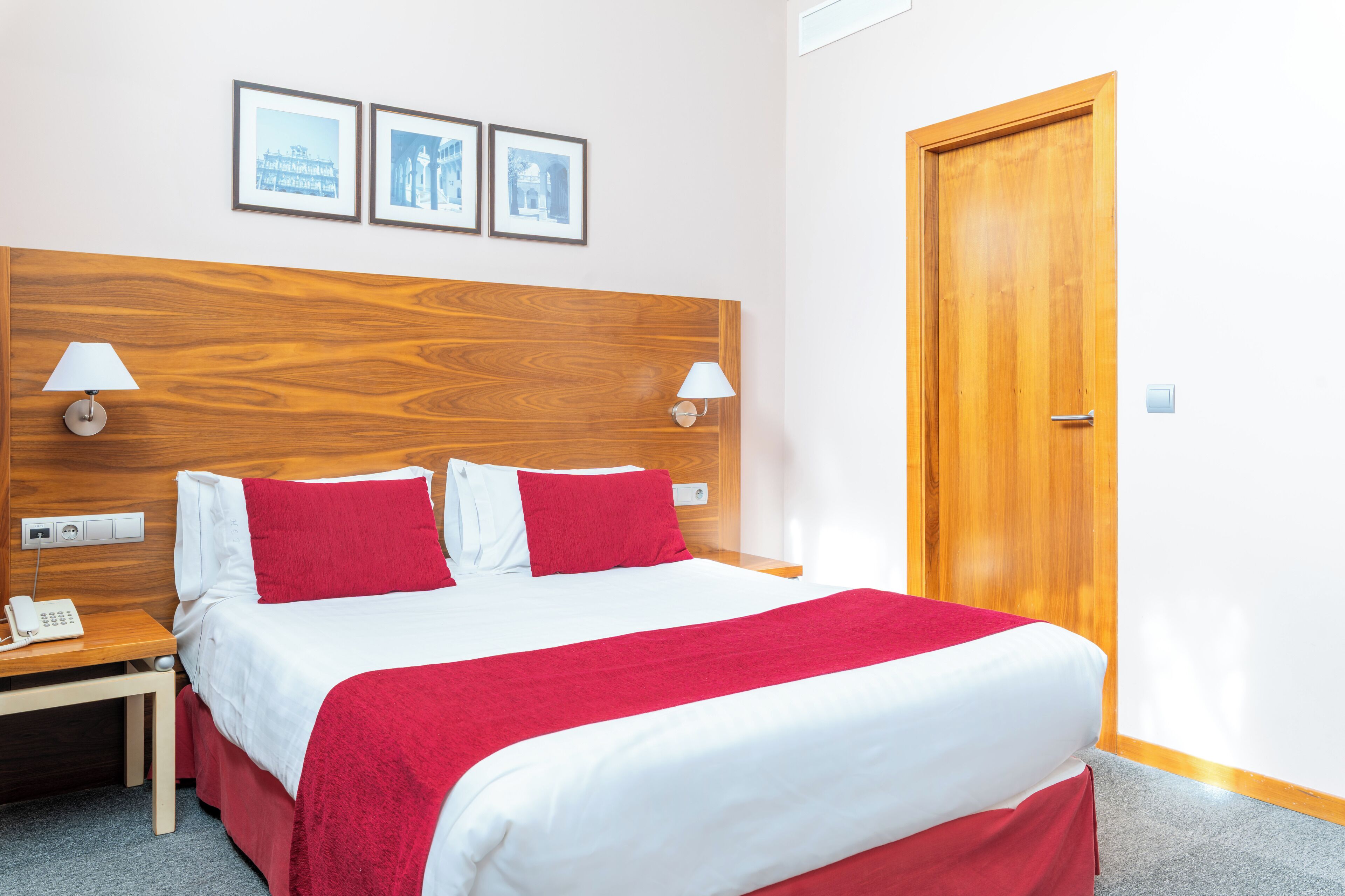 A hotel room with a wooden headboard, a bed with white and red bedding, two bedside lamps, a nightstand with a phone, and a door.