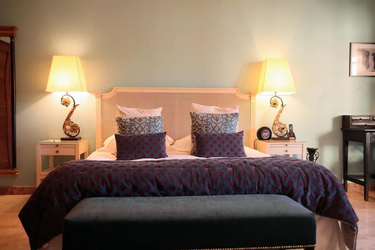 Een net opgemaakt bed met gestreepte kussens, twee nachtlampjes en een nachtkastje met een klok en een vaas.