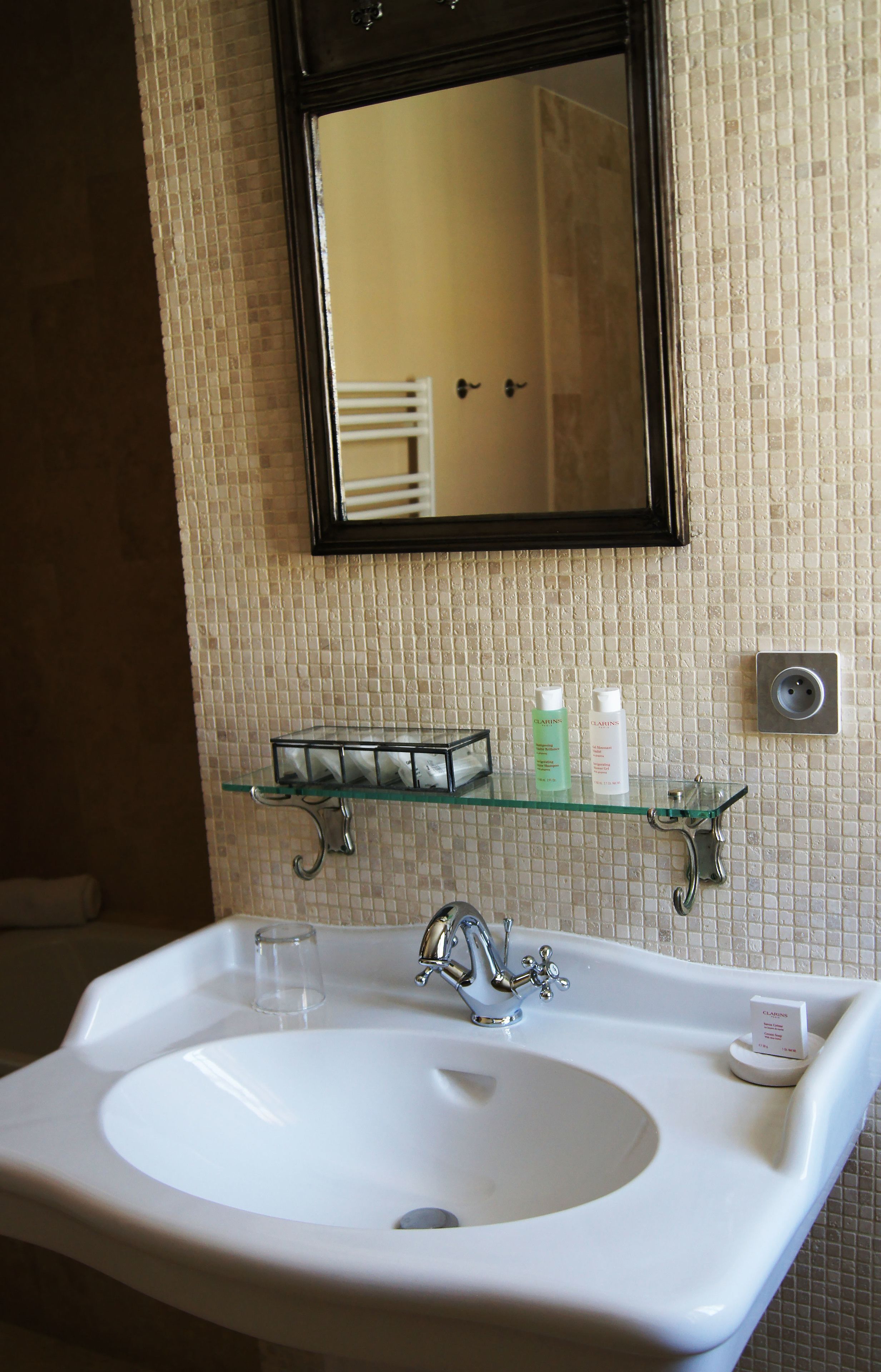 Une salle de bain avec un lavabo blanc, une étagère en verre où sont rangés les articles de toilette, un miroir et un mur recouvert de carreaux.
