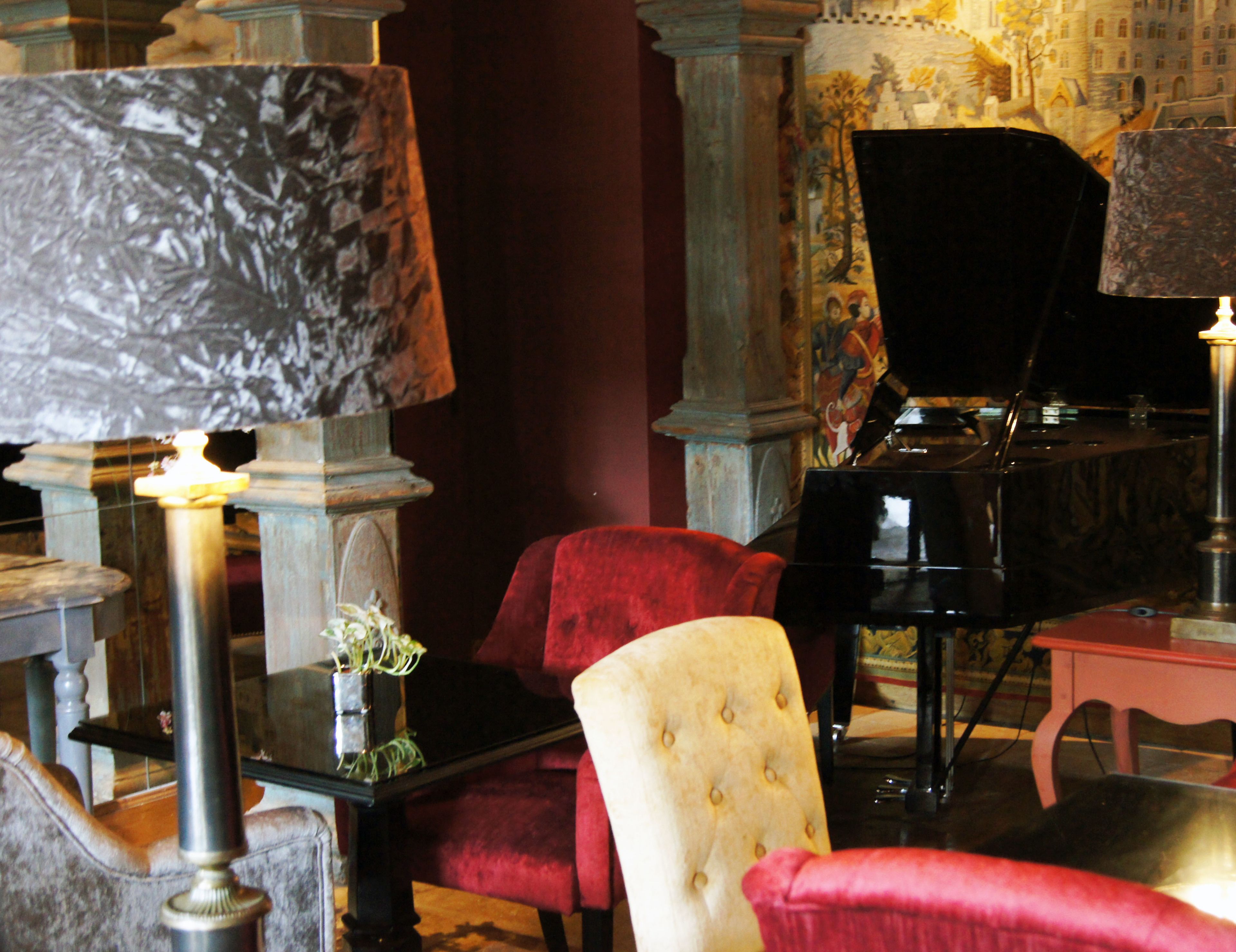 A room with a grand piano, a red velvet armchair, a white tufted chair, and a table with a lamp.