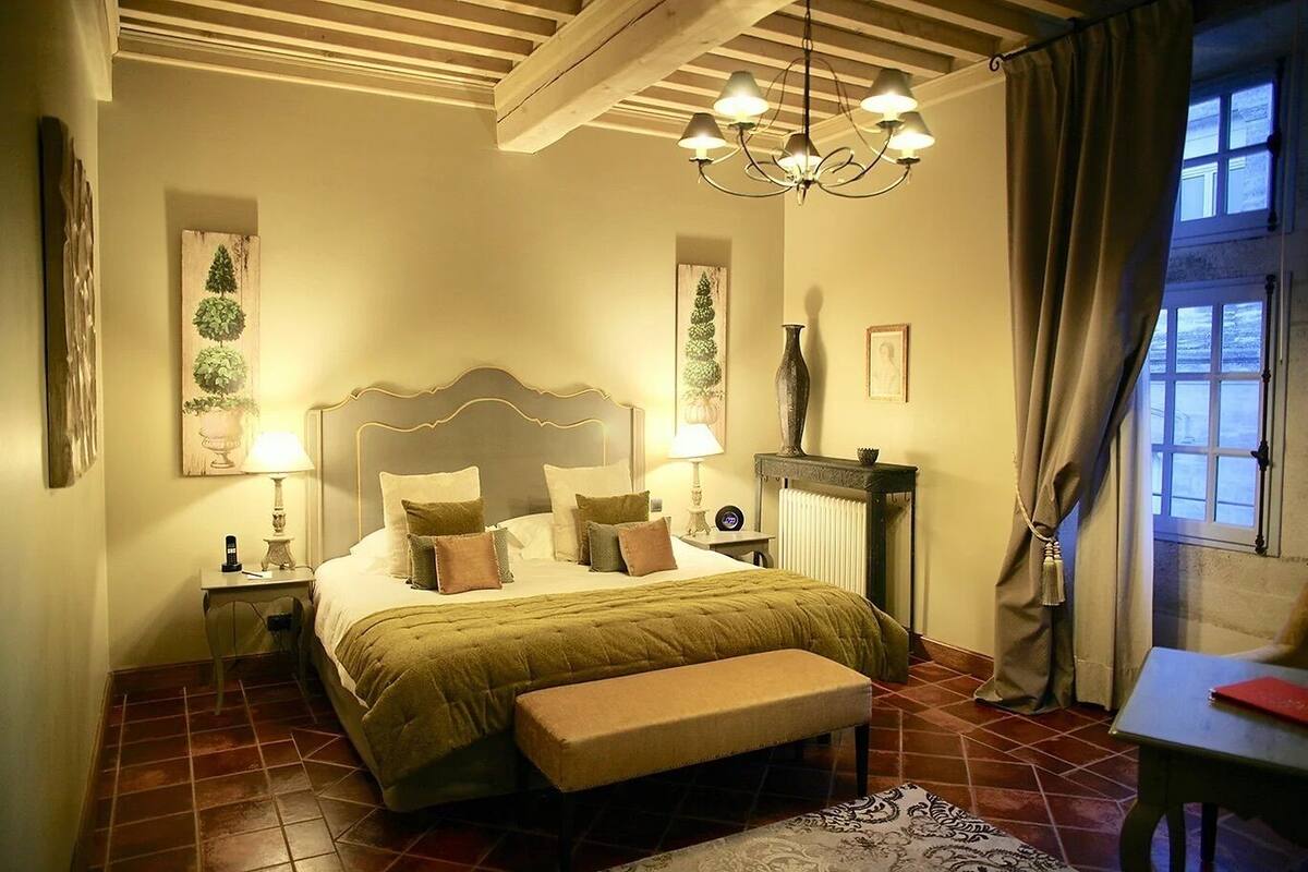 Een slaapkamer met een groot bed, nachtkastjes, een bankje, een kroonluchter en uitzicht op het omliggende landschap door een raam.