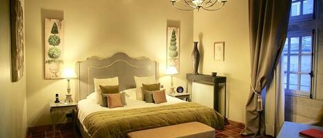 A bedroom with a large bed, bedside tables, a bench, a chandelier, and a view of the outside through a window.