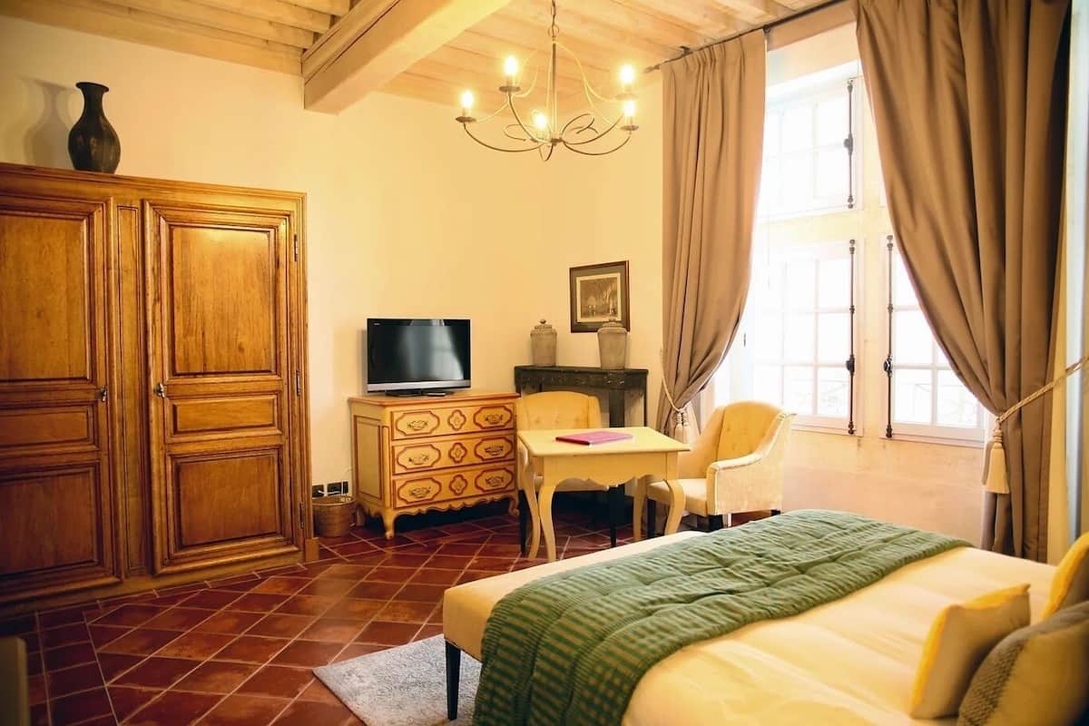 Een hotelkamer met een houten garderobe, een televisie op een houten kast, een bed met een groene deken, een klein tafeltje met een stoel, en een raam met gordijnen.
