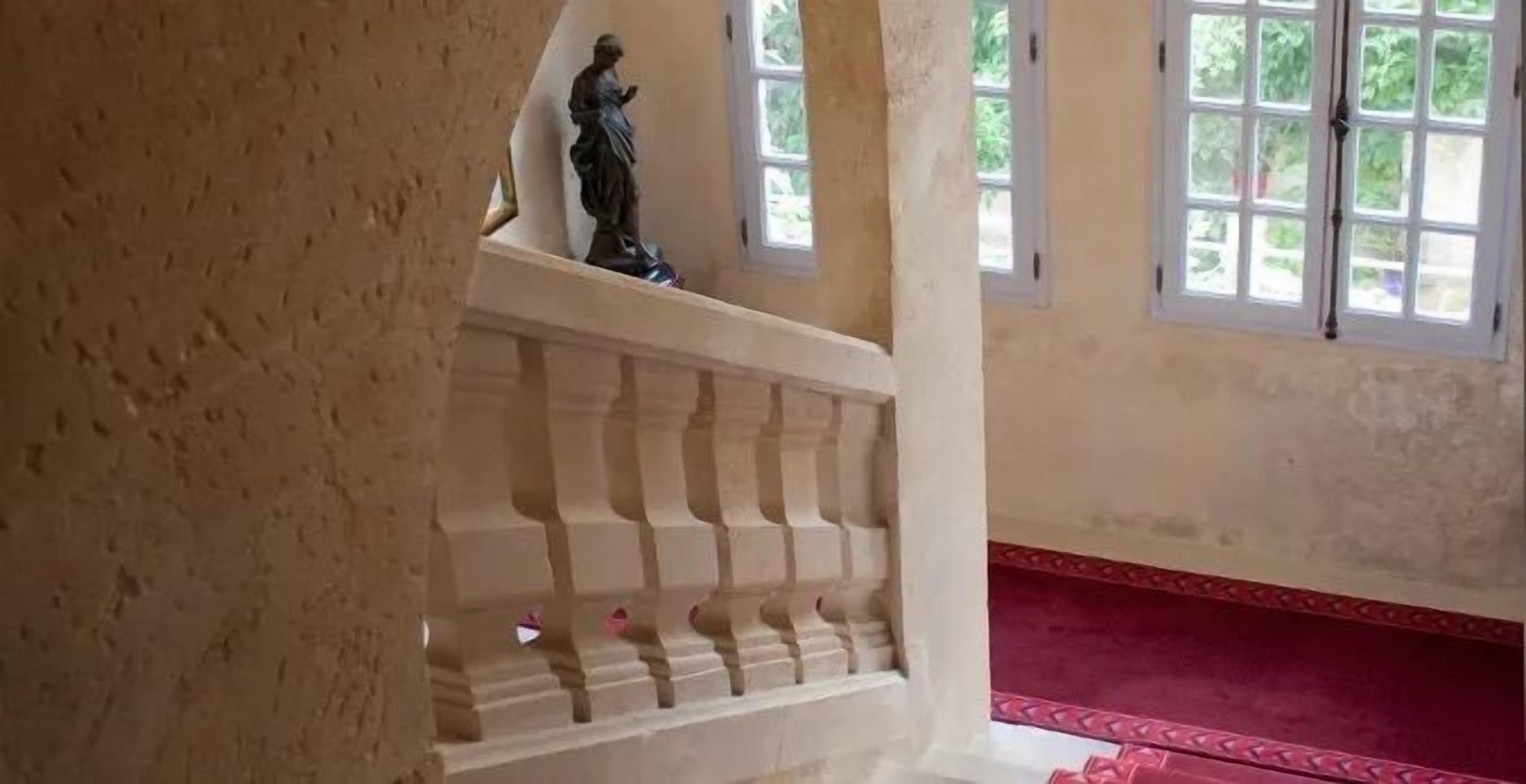 Un escalier orné d’une statue sur la plateforme, une zone recouverte d’un tapis rouge et de grandes fenêtres donnant sur un paysage verdoyant.