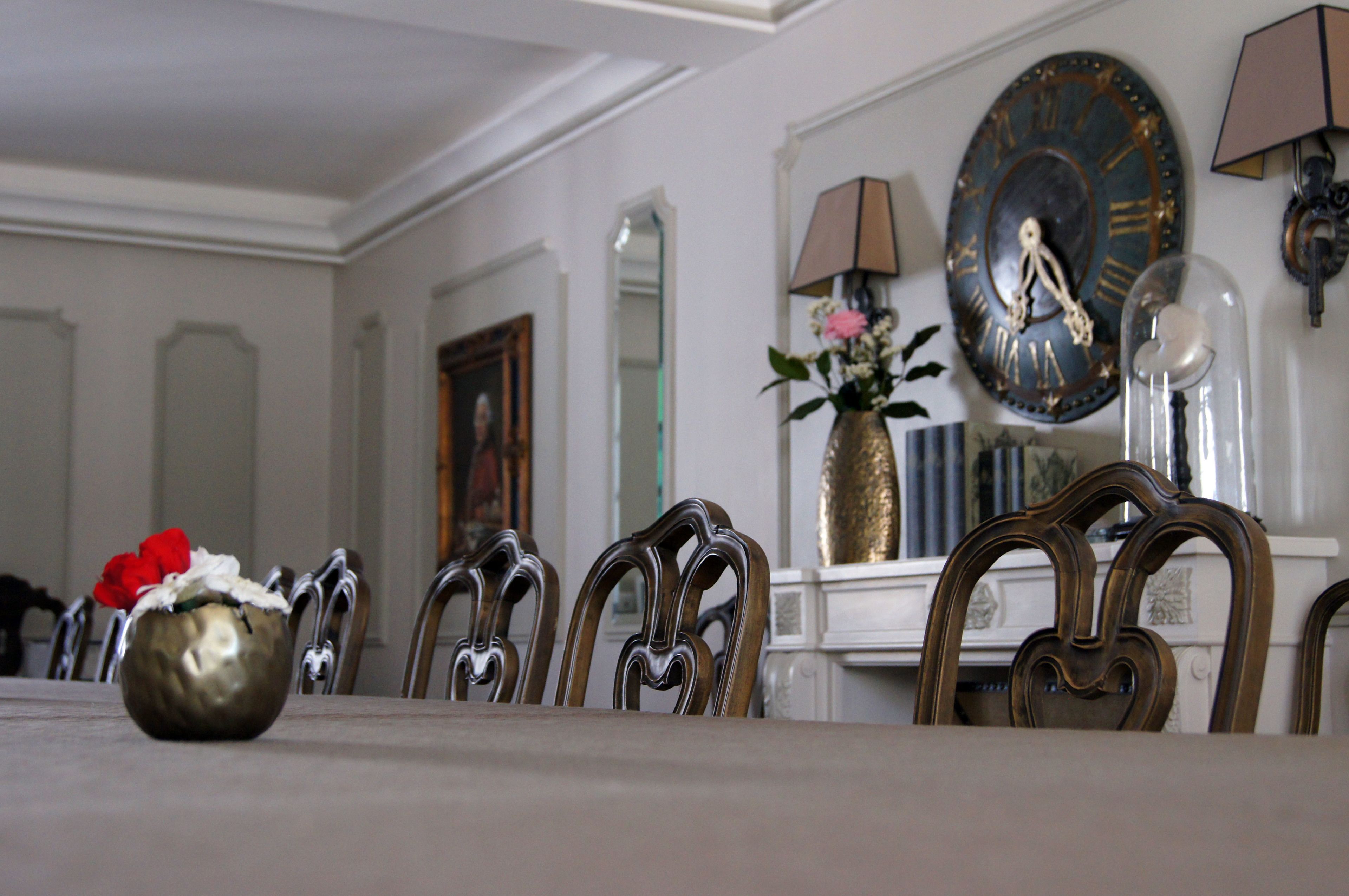 Un comedor con una mesa dispuesta para cuatro personas, rodeada de sillas ornamentales. Hay un jarrón con flores sobre la mesa, además de un reloj de pared y una lámpara en la pared.