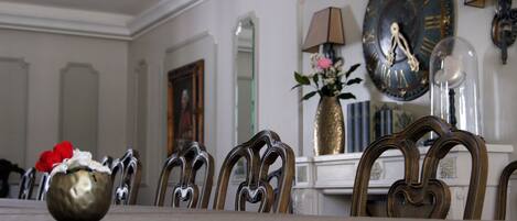 Un comedor con una mesa dispuesta para cuatro personas, rodeada de sillas ornamentales. Hay un jarrón con flores sobre la mesa, además de un reloj de pared y una lámpara en la pared.