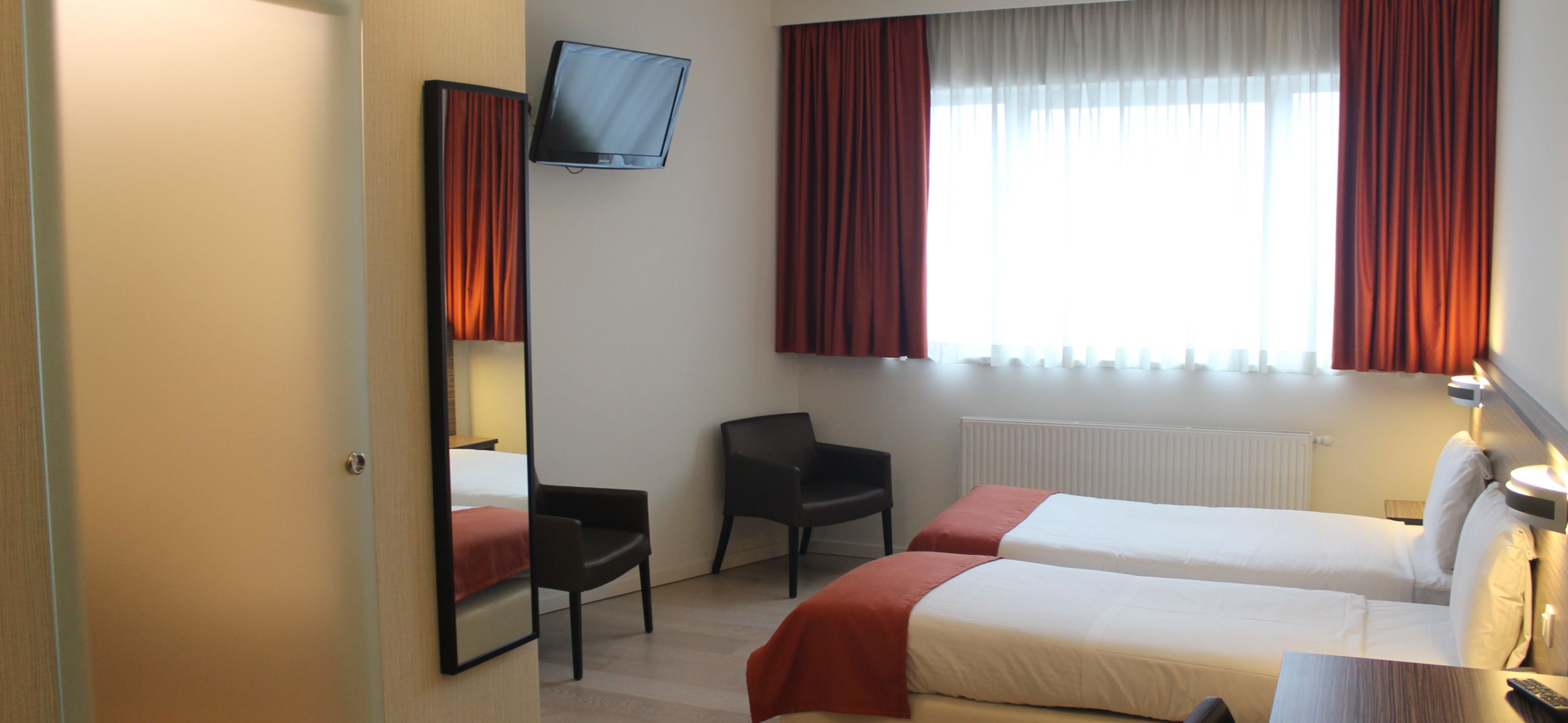 A hotel room with two beds, a TV mounted on the wall, a chair, and a desk with a lamp.