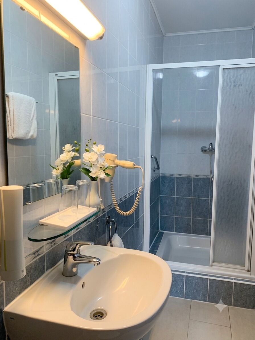 A bathroom with a white sink, a glass shelf with toiletries, a handheld hair dryer, and a shower enclosure.