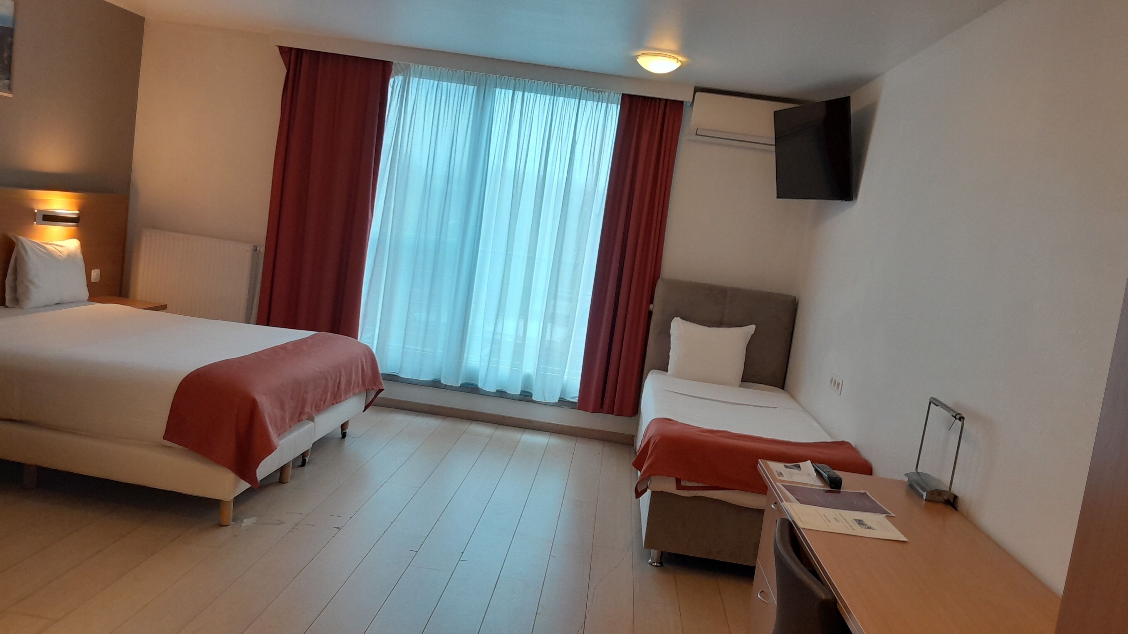 Quarto de hotel com duas camas, uma escrivaninha, uma cadeira, televisão fixada na parede e uma janela com cortinas.