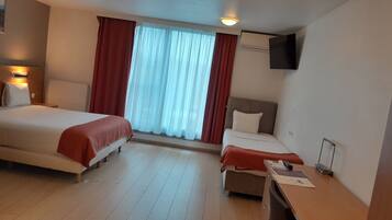 Quarto de hotel com duas camas, uma escrivaninha, uma cadeira, televisão fixada na parede e uma janela com cortinas.