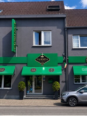 A hotel with a green awning and a sign that reads 'TAORMINA HOTEL/RESTAURANT.' There is a car parked in front, and the building has a grey facade with a tiled roof.