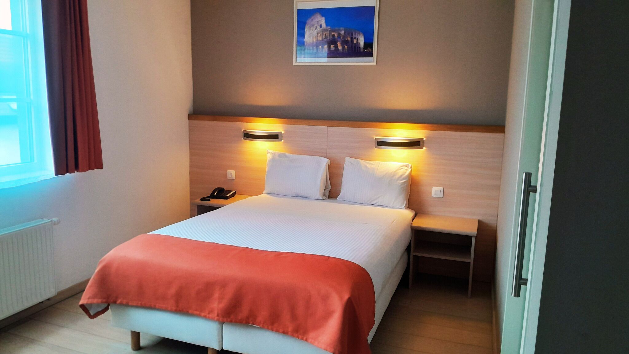A hotel room with a bed, bedside tables, a nightstand, a telephone, and a framed picture on the wall.