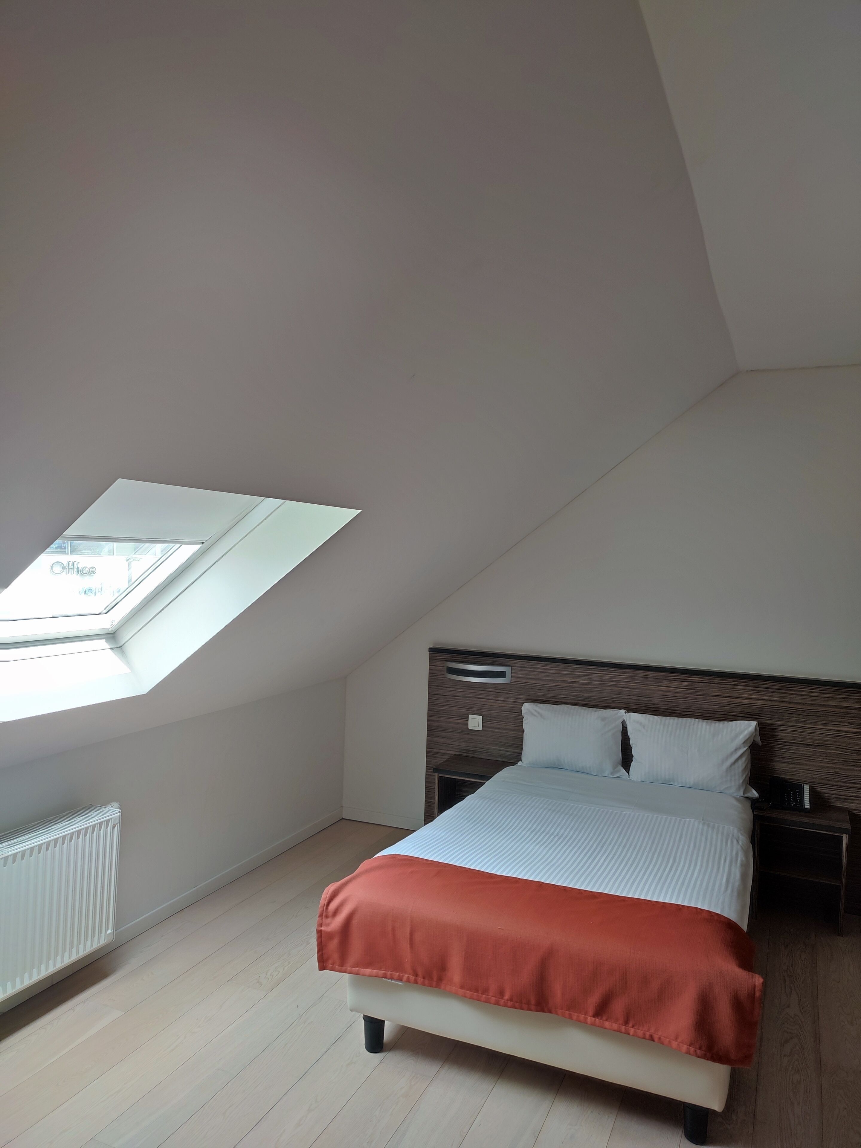 Un dormitorio con tragaluz, una cama con manta roja, una mesita de noche y un radiador.