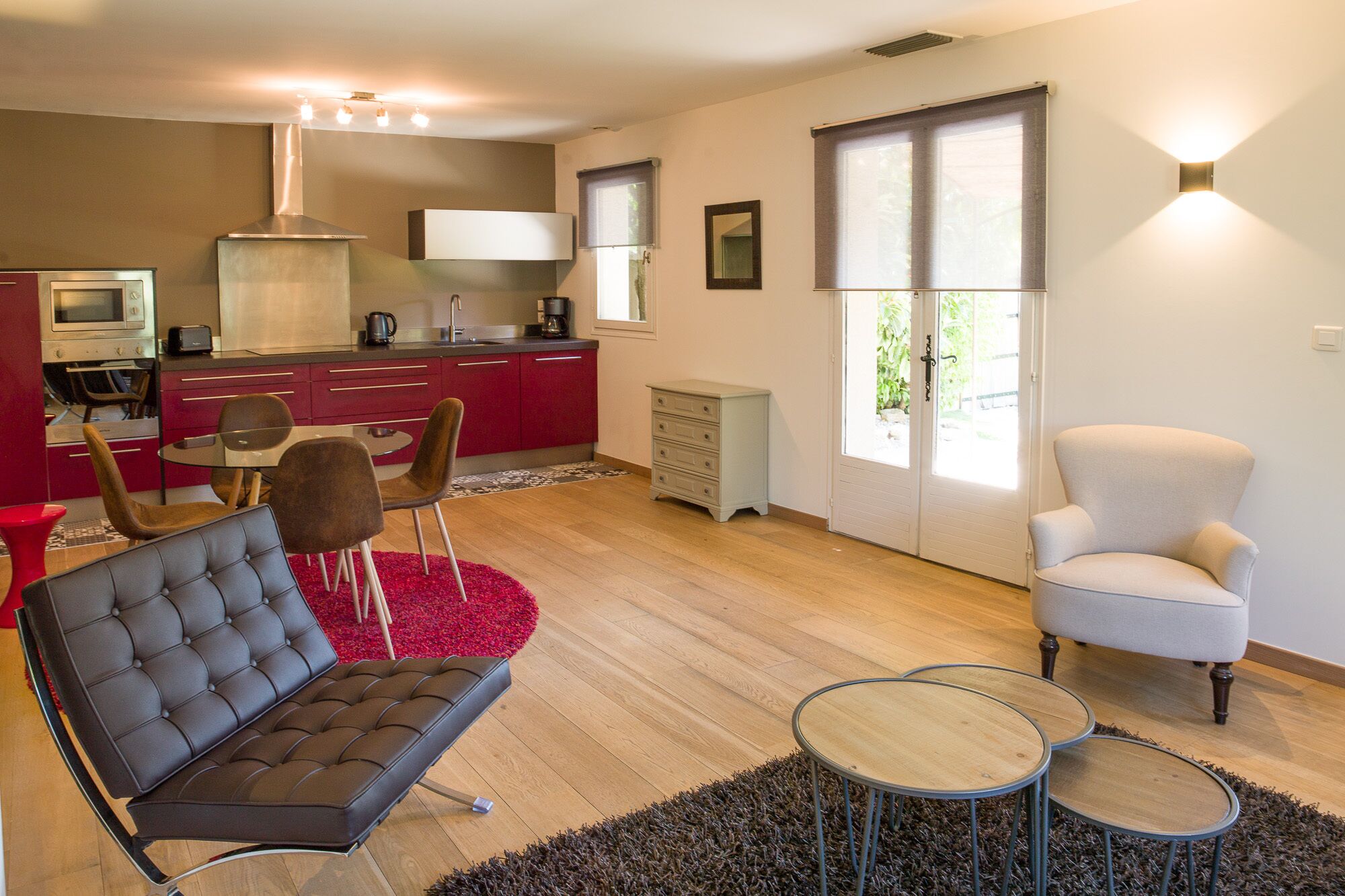 A modern living room with a sofa, armchair, and coffee table, a kitchen area with red cabinets, and a large window providing natural light.