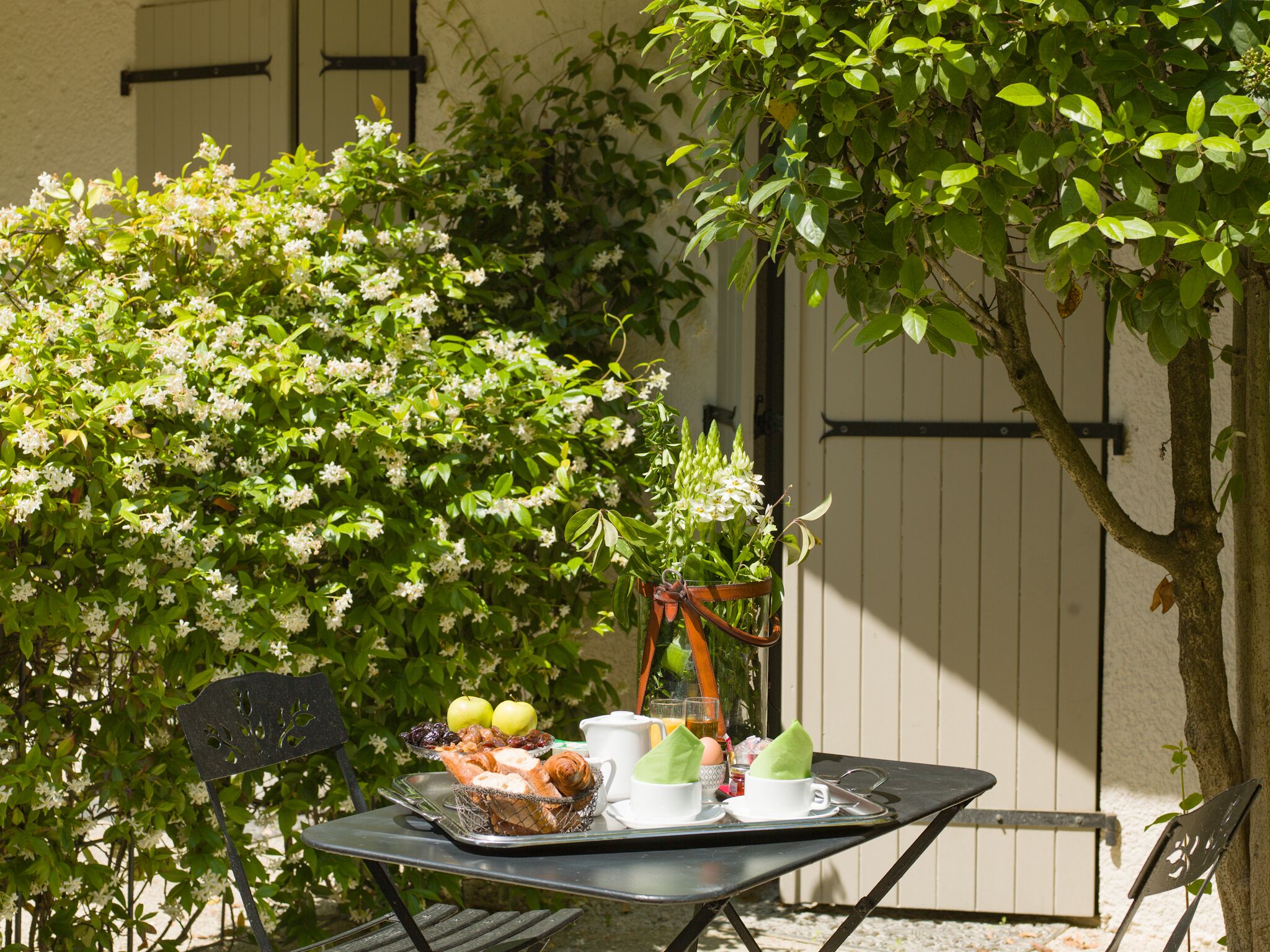 A table set outdoors with a basket of bread, apples, and a cup of tea, surrounded by greenery and a white wall.