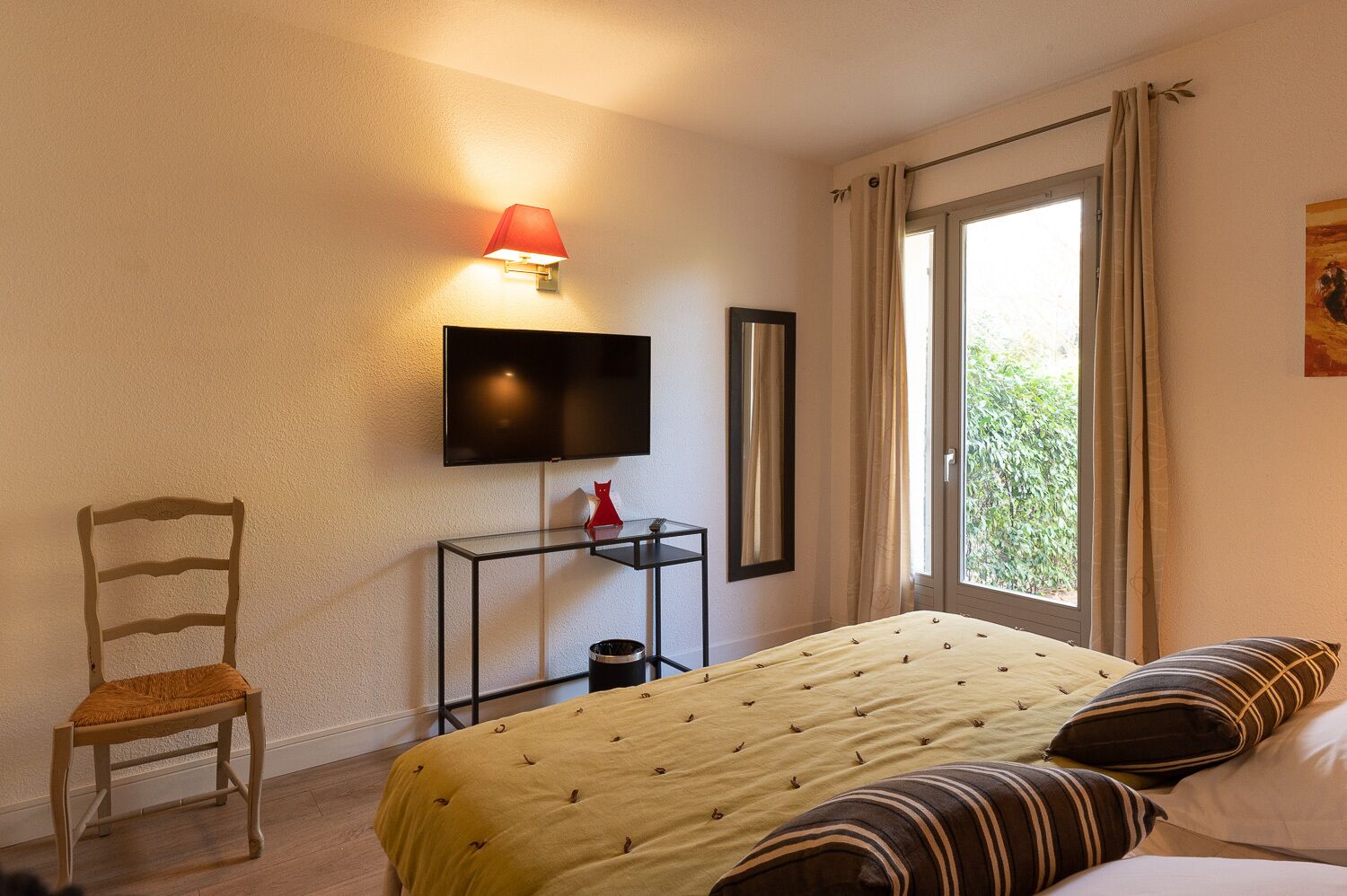 A hotel room with a bed, a TV mounted on the wall, a chair, a small table with a lamp, and a window with curtains.
