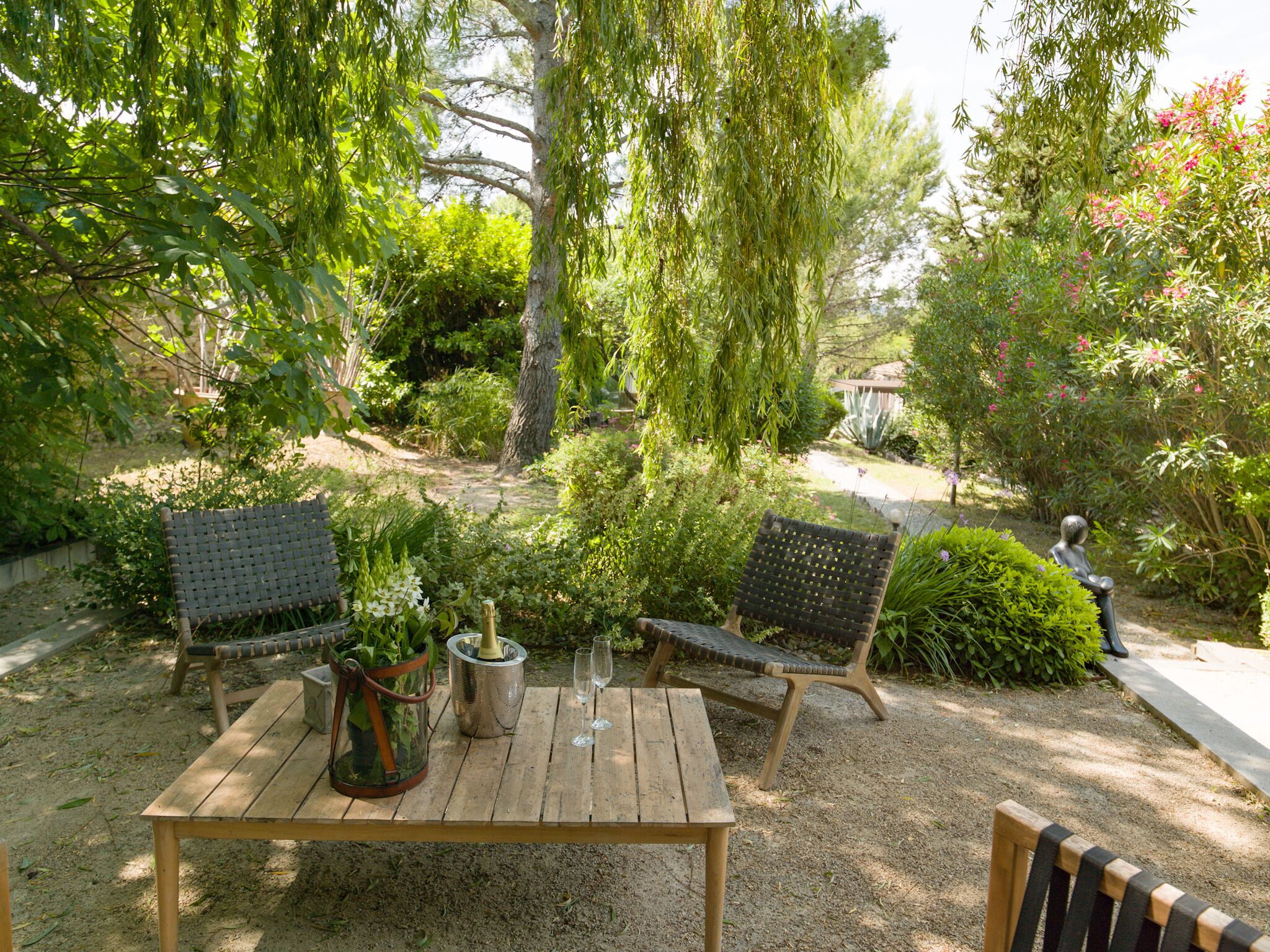 An outdoor seating area with a wooden table and two chairs, surrounded by lush greenery and a statue.