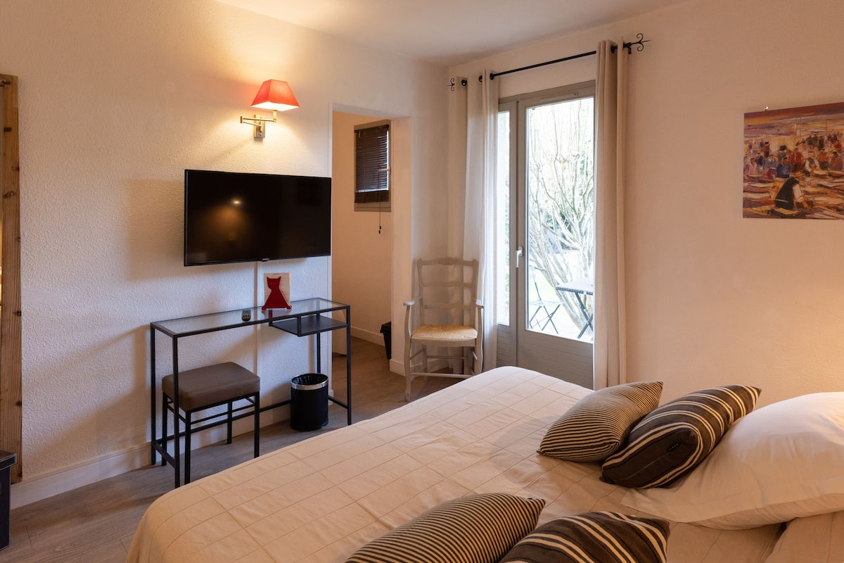 A hotel room with a bed, a desk with a chair, a TV mounted on the wall, and a balcony with a view of trees.
