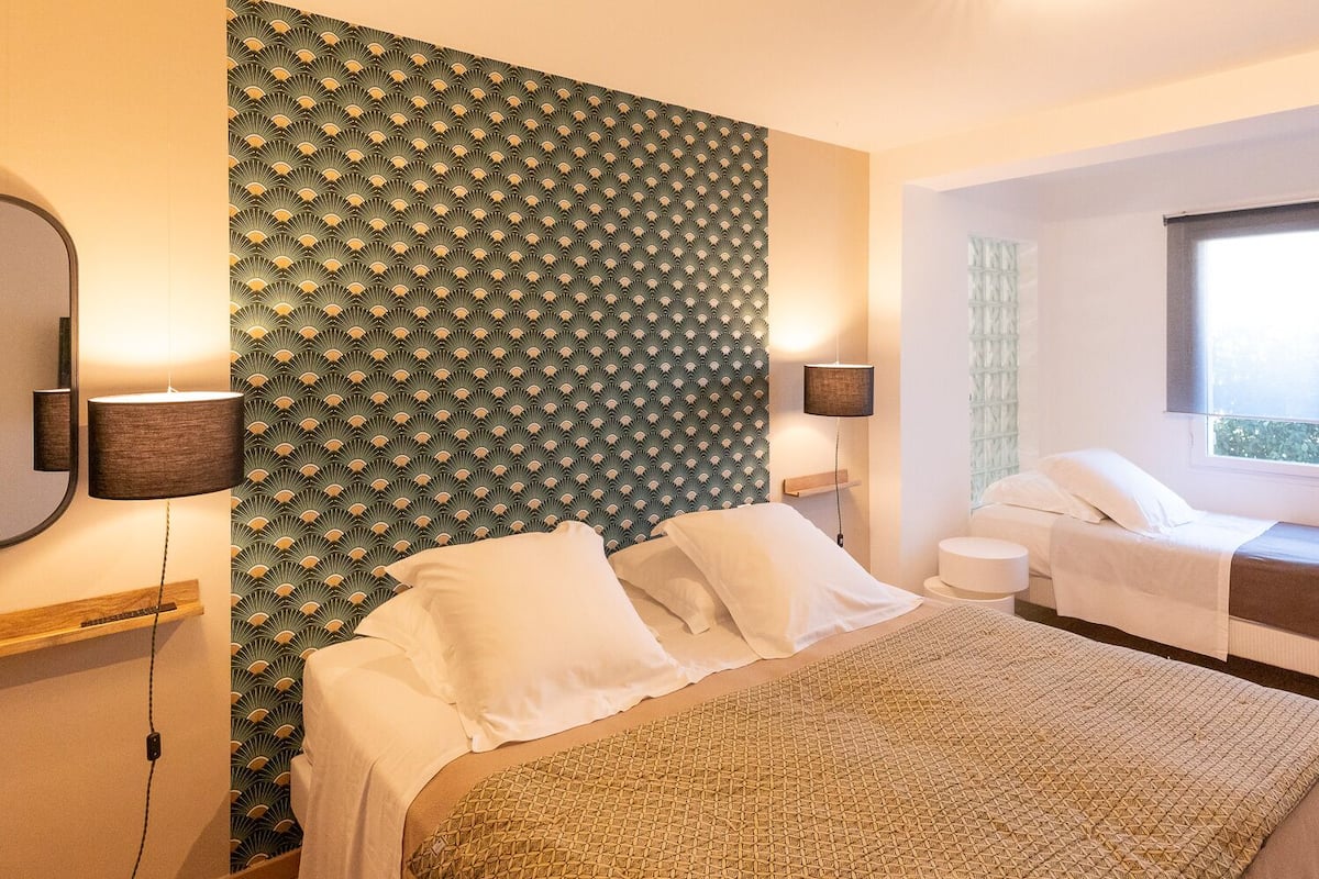 A hotel room with a bed, bedside tables, a mirror, a wall with a geometric pattern, a window, and a bedside lamp.