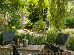 An outdoor seating area with a wooden table and two chairs, surrounded by lush greenery and a weeping willow tree. - Hotel de Charme L'Amandière (St.-Rémy-de-Provence)