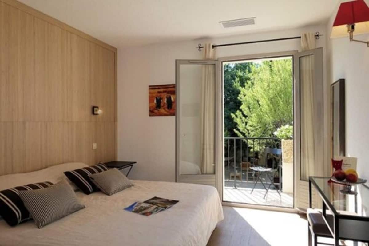A bedroom with a bed, a small table with a lamp, and a balcony with a view of trees.