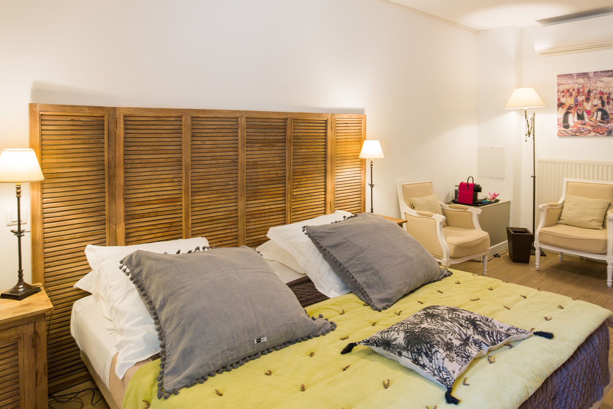 Una habitación de hotel con cabecera de madera, dos camas con ropa de cama gris y blanca, un edredón amarillo y una mesita con un bolso rojo.