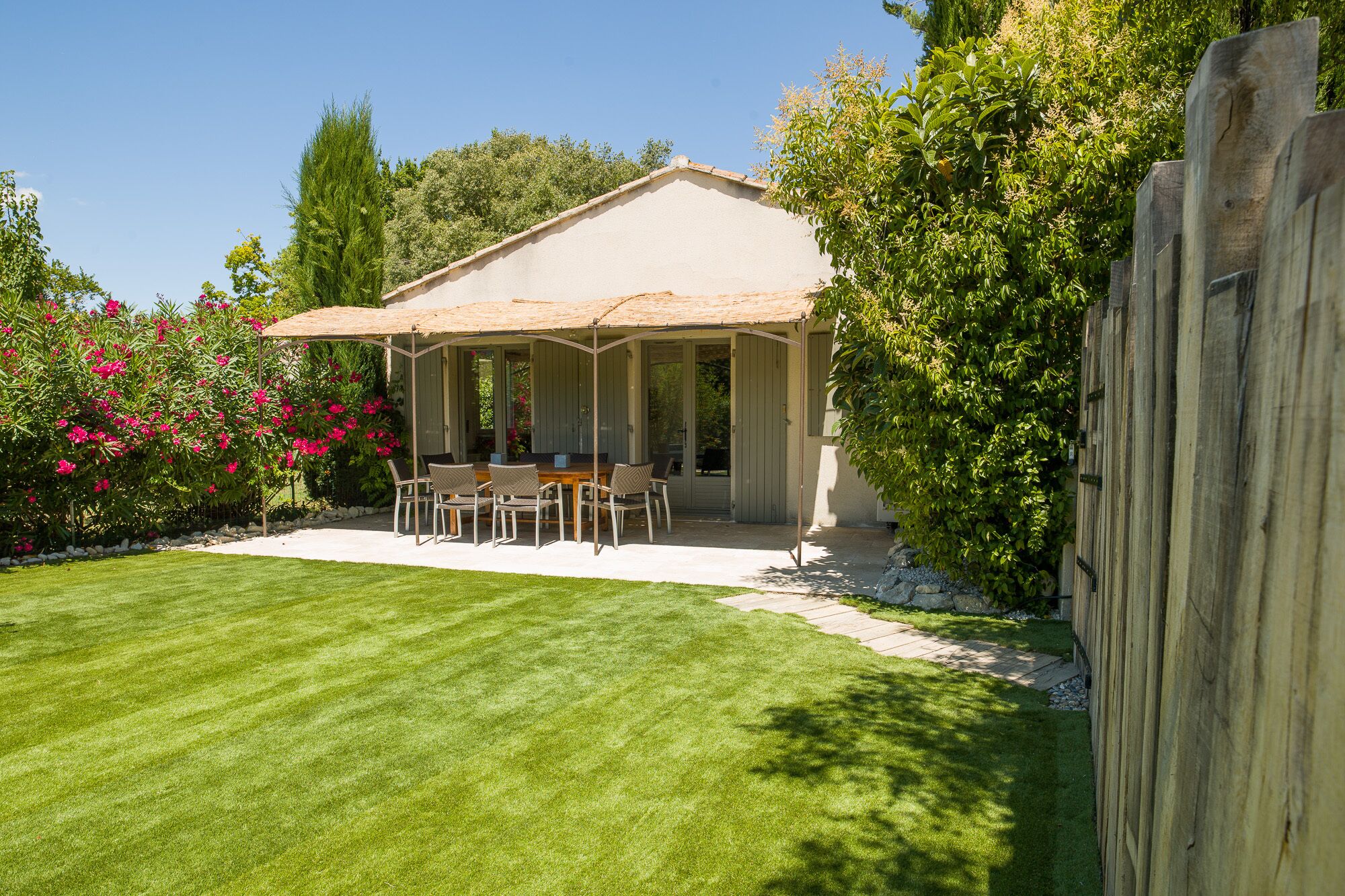 A covered patio with a table and chairs, surrounded by a well-maintained lawn and flowering bushes.