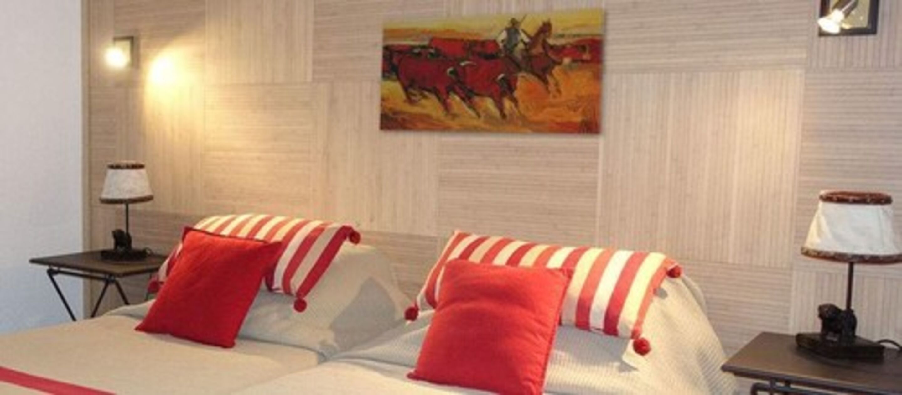Habitación de hotel con dos camas, cada una con almohadas a rayas rojas y blancas, un cuadro de una carrera de caballos en la pared y una lámpara a cada lado de la cama.