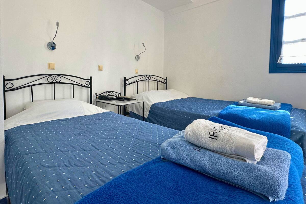 Habitación de hotel con dos camas, cada una con cubrecama azul y almohadas blancas, y una ventana con marco azul.
