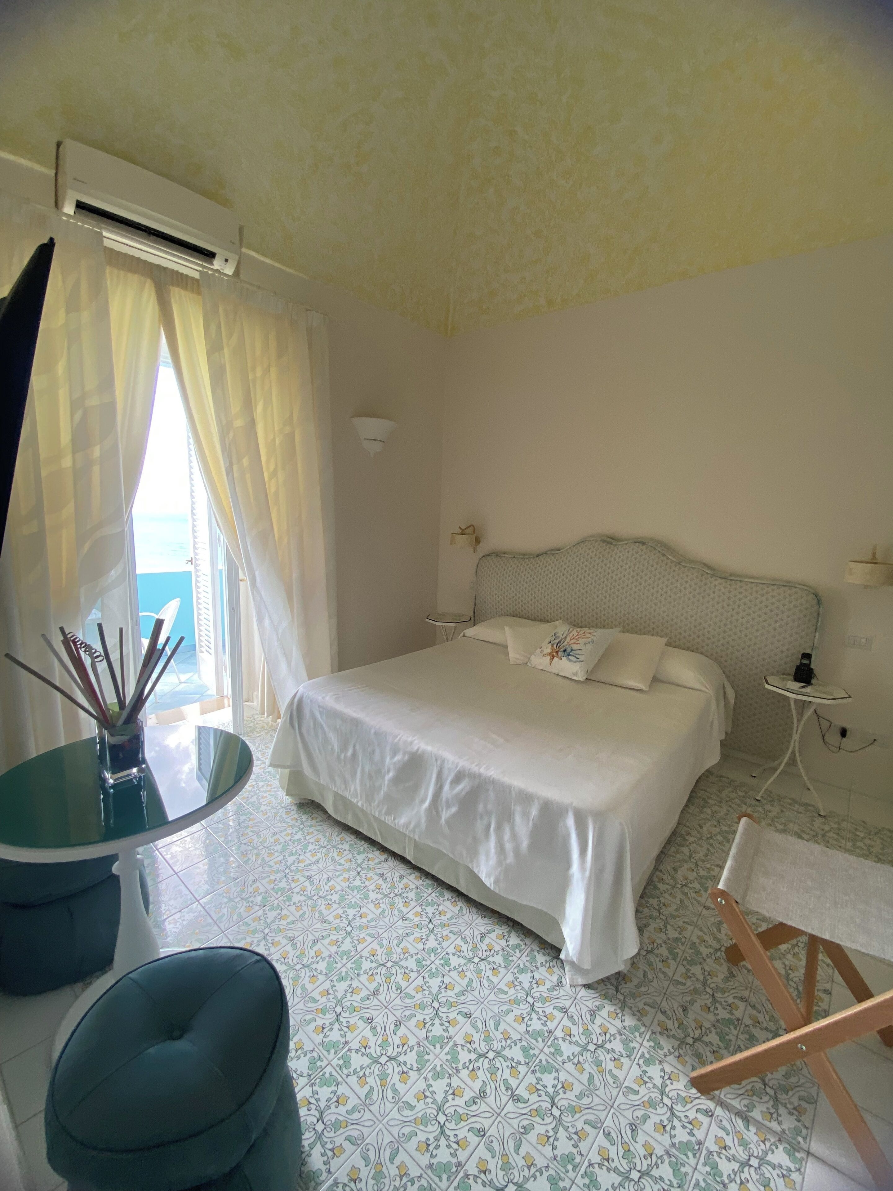 A hotel room with a bed, a small table, a chair, and a view of the sea through a window.