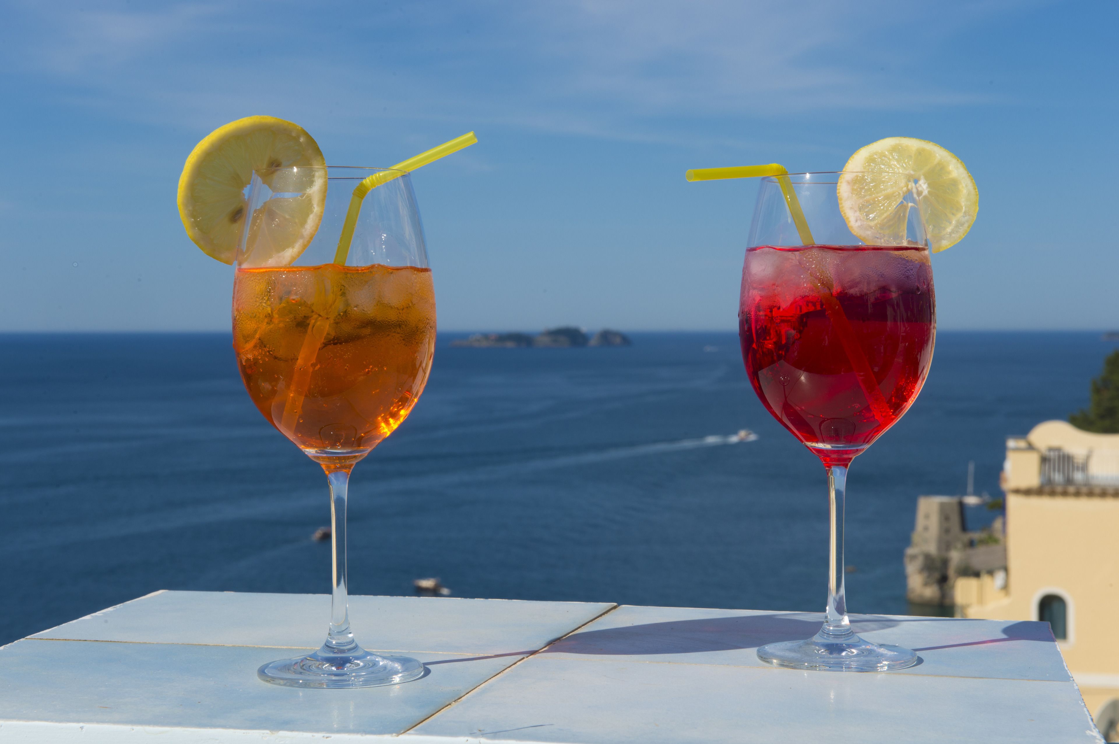 Two glasses of beverages with lemon slices and straws on a table with a view of the sea and buildings in the background.