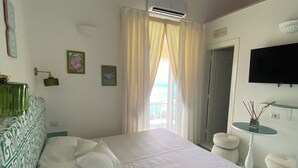 A hotel room with a large bed, a small table with a vase, a mounted TV, and a window with curtains. - Palazzo Talamo (Positano)