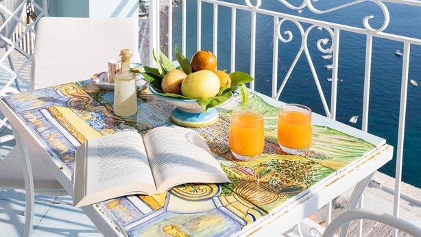 A table with a book, a bottle, a bowl of fruit, and two glasses of orange juice, set on a balcony with a view of the sea and mountains. - Palazzo Talamo (Positano)