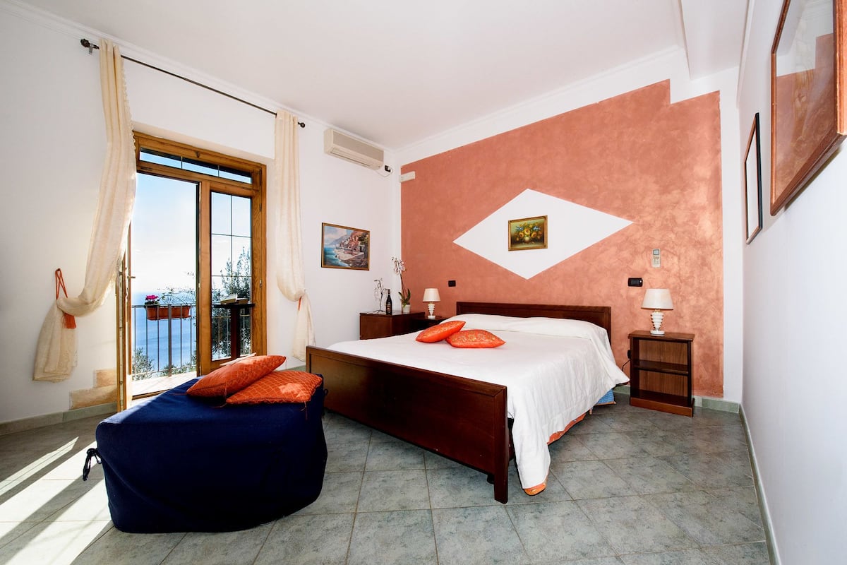 A bedroom with a bed, a nightstand, a chair, a window with curtains, and a wall with a picture.
