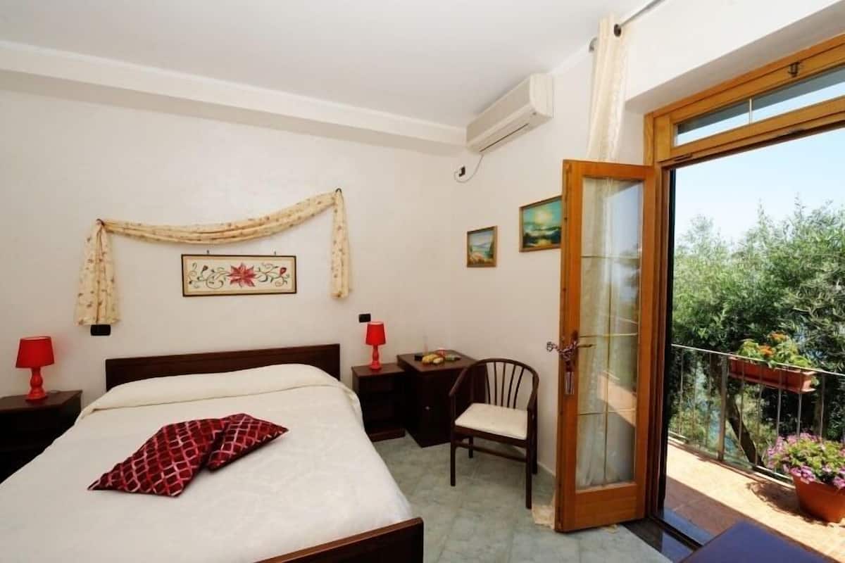 A bedroom with a bed, a chair, a nightstand, a table, and a balcony with plants.