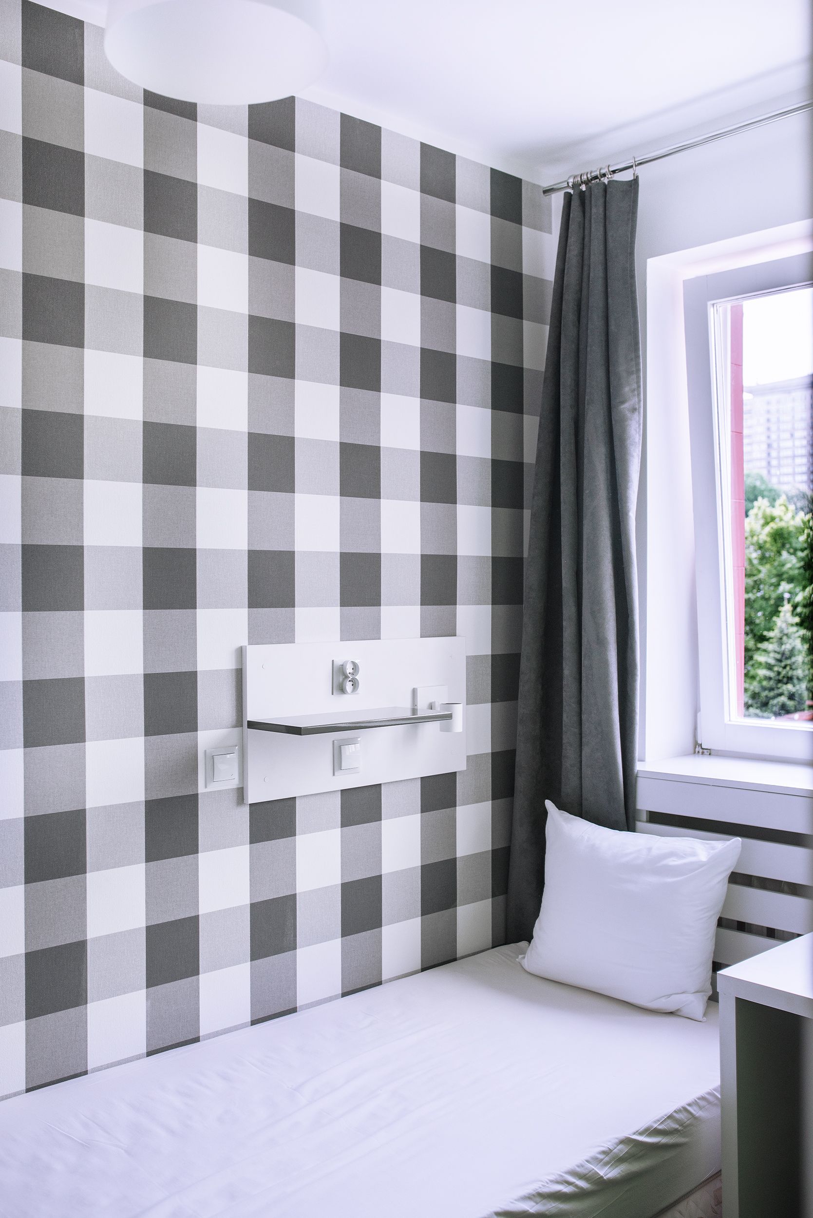 A small, neatly arranged bedroom with a checkered wall pattern, a window with a view, and a bedside table with a lamp.