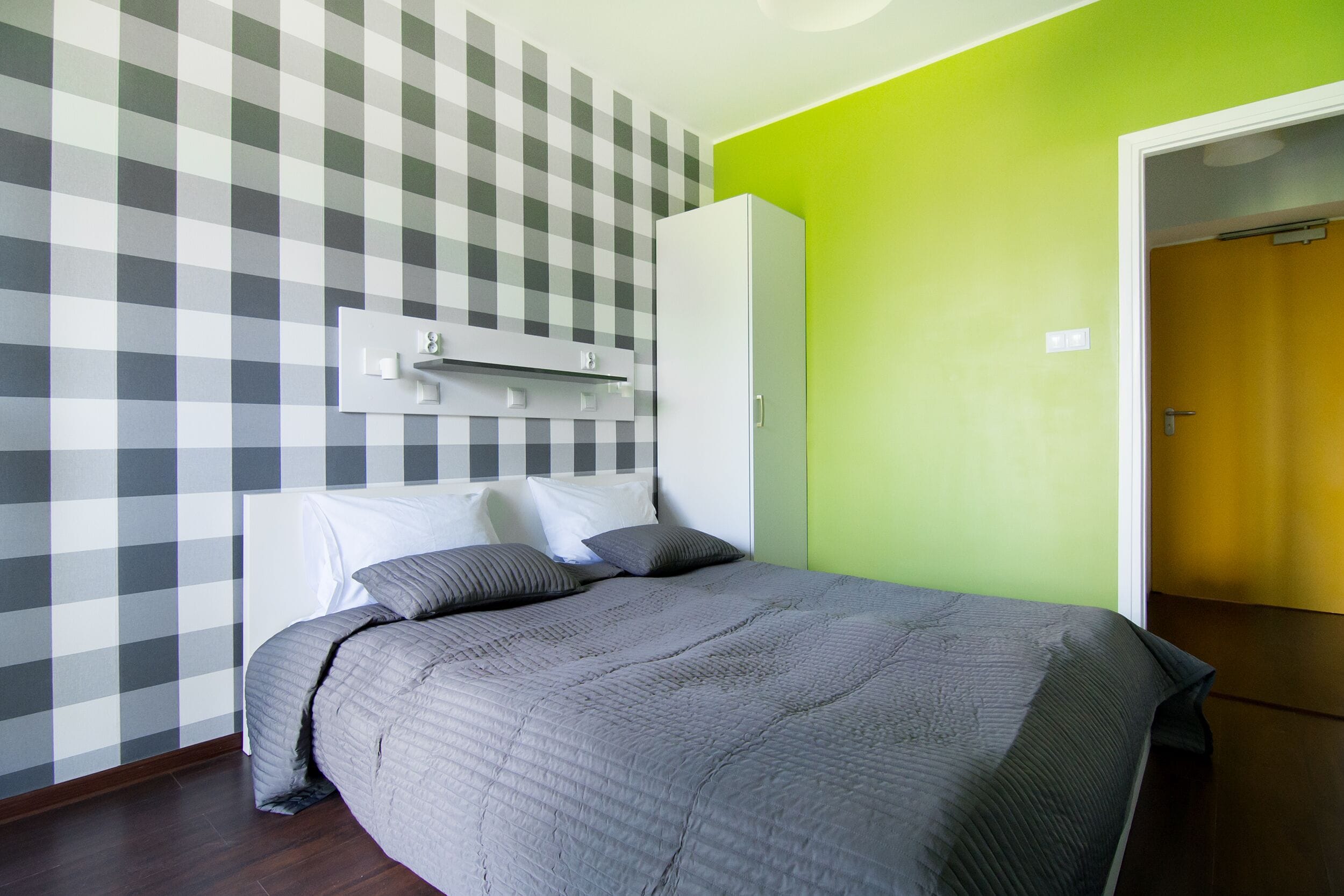 A bedroom with a checkered wall, a bed with grey bedding, a wardrobe, and a door to another room.