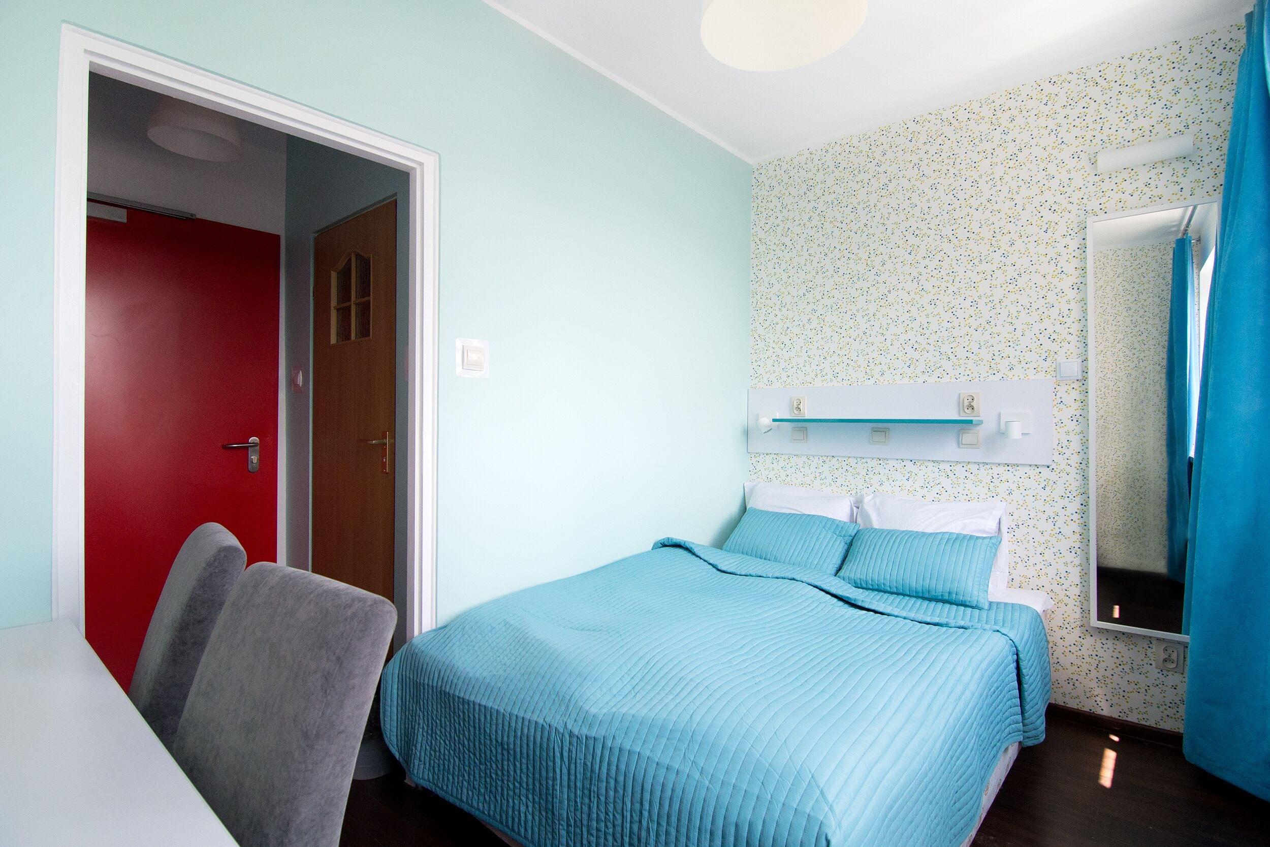 A room with a bed, a desk, and a chair. There is a door and a mirror on the wall.