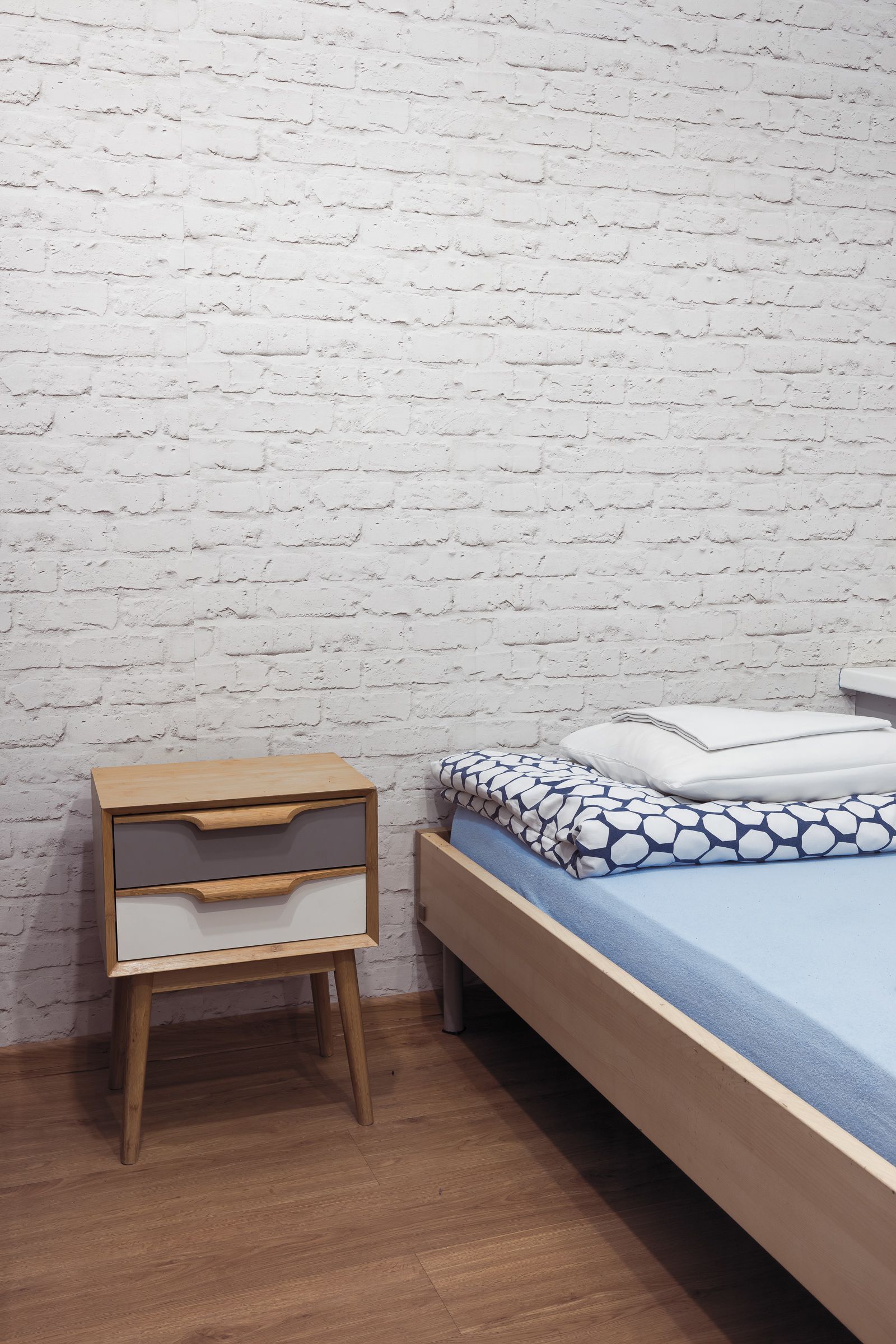 Un dormitorio con una mesita de noche de madera, una cama con colchón azul y almohadas estampadas, y una pared de ladrillo blanco.