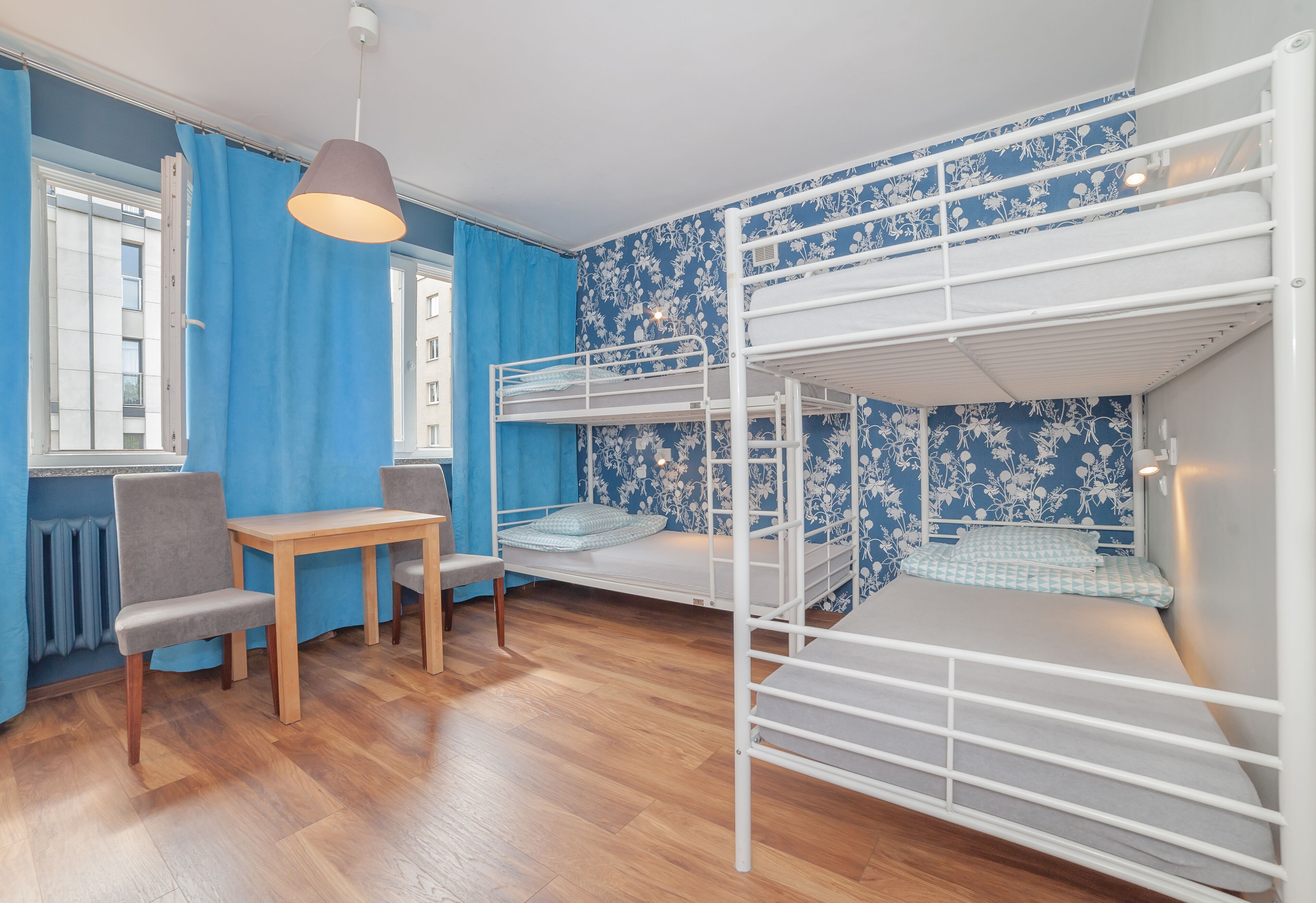 A bunk bed room with a wooden table and chairs, a window with blue curtains, and a patterned wall.