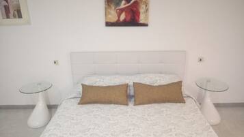 A bedroom with a white bedspread, two brown pillows, a white headboard, and a glass table on each side.