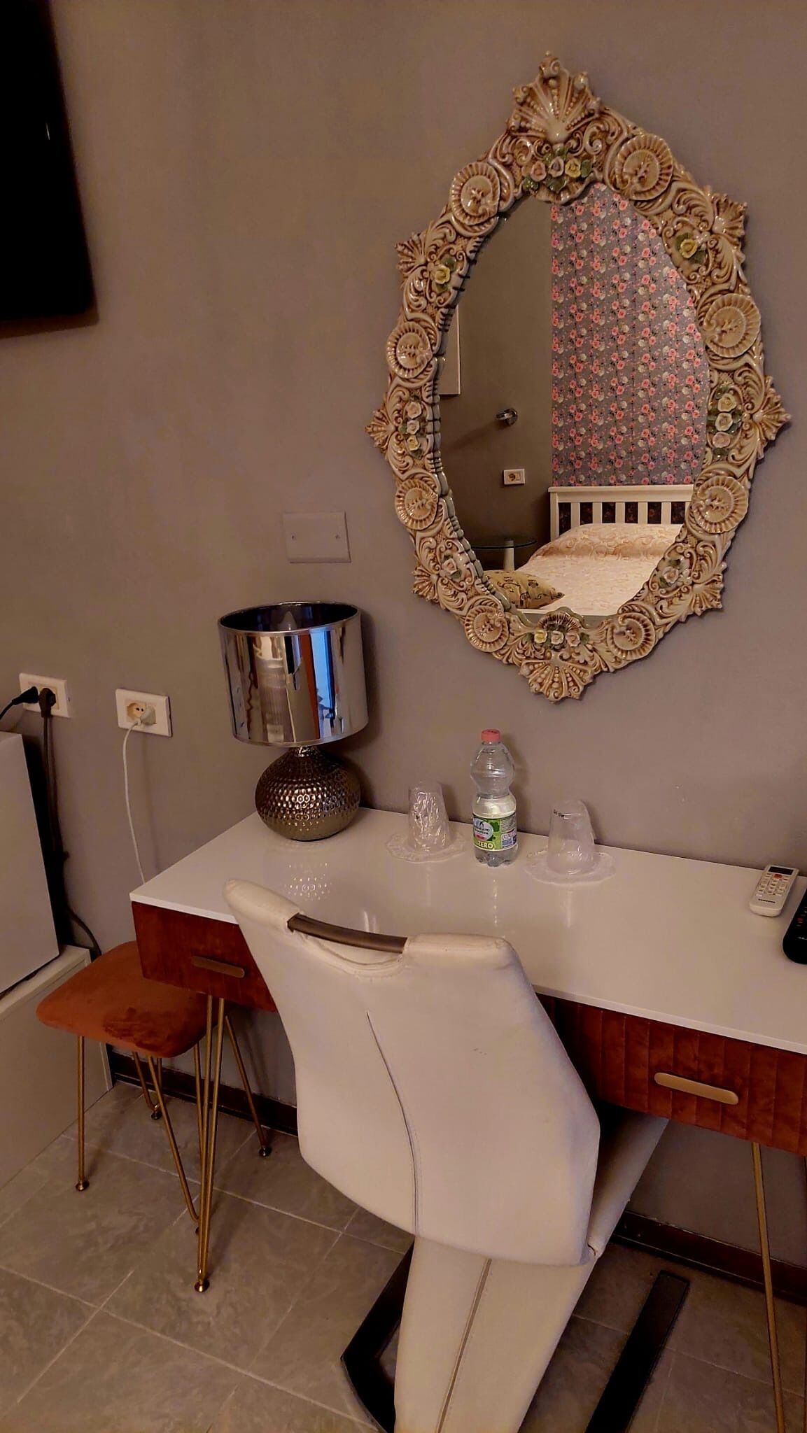 A hotel room with a white vanity, a large ornate mirror, a small stool, a metallic lamp, and a bottle of water.