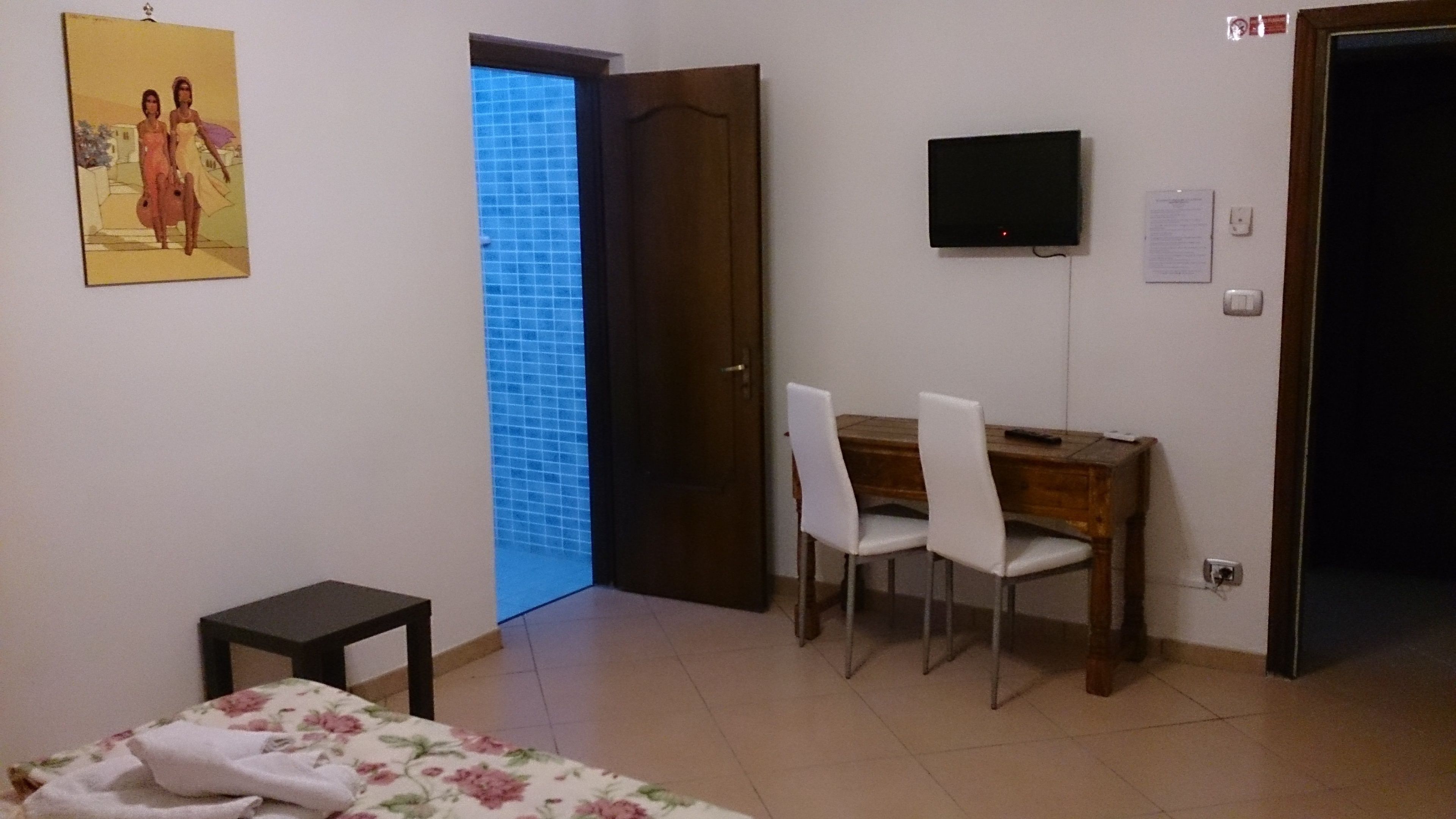 A room with a bed, a desk with chairs, a television, and a door leading to a balcony.