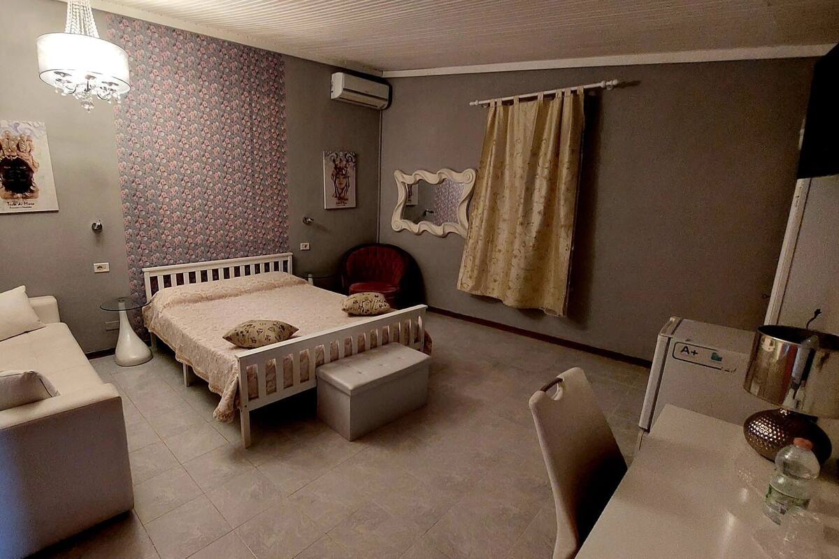 A bedroom with a bed, a sofa, a chair, a small table, a mirror, and an air conditioning unit.
