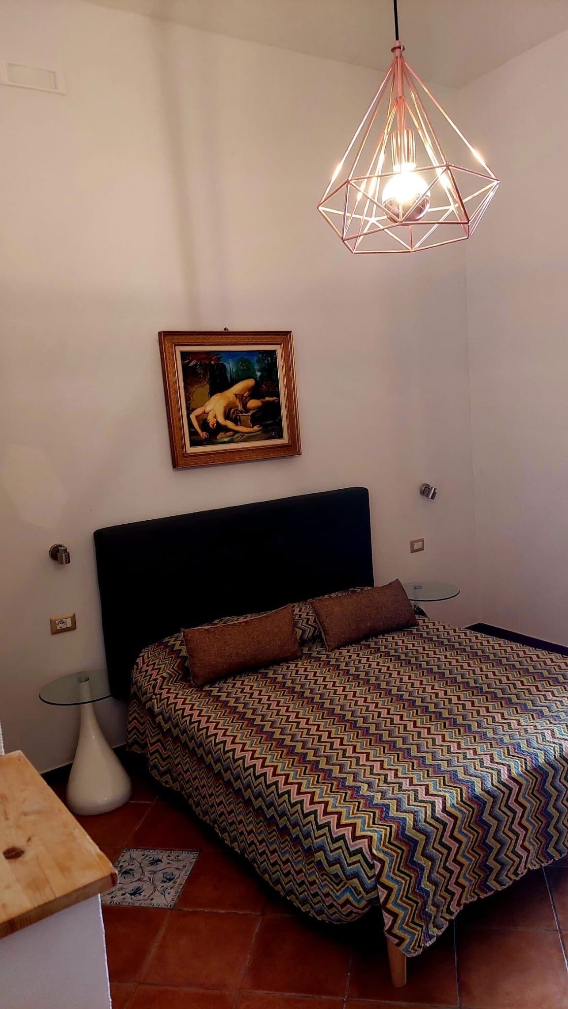 Un dormitorio con colcha estampada, cabecero negro, un cuadro enmarcado, lámpara colgante y una mesita de noche.
