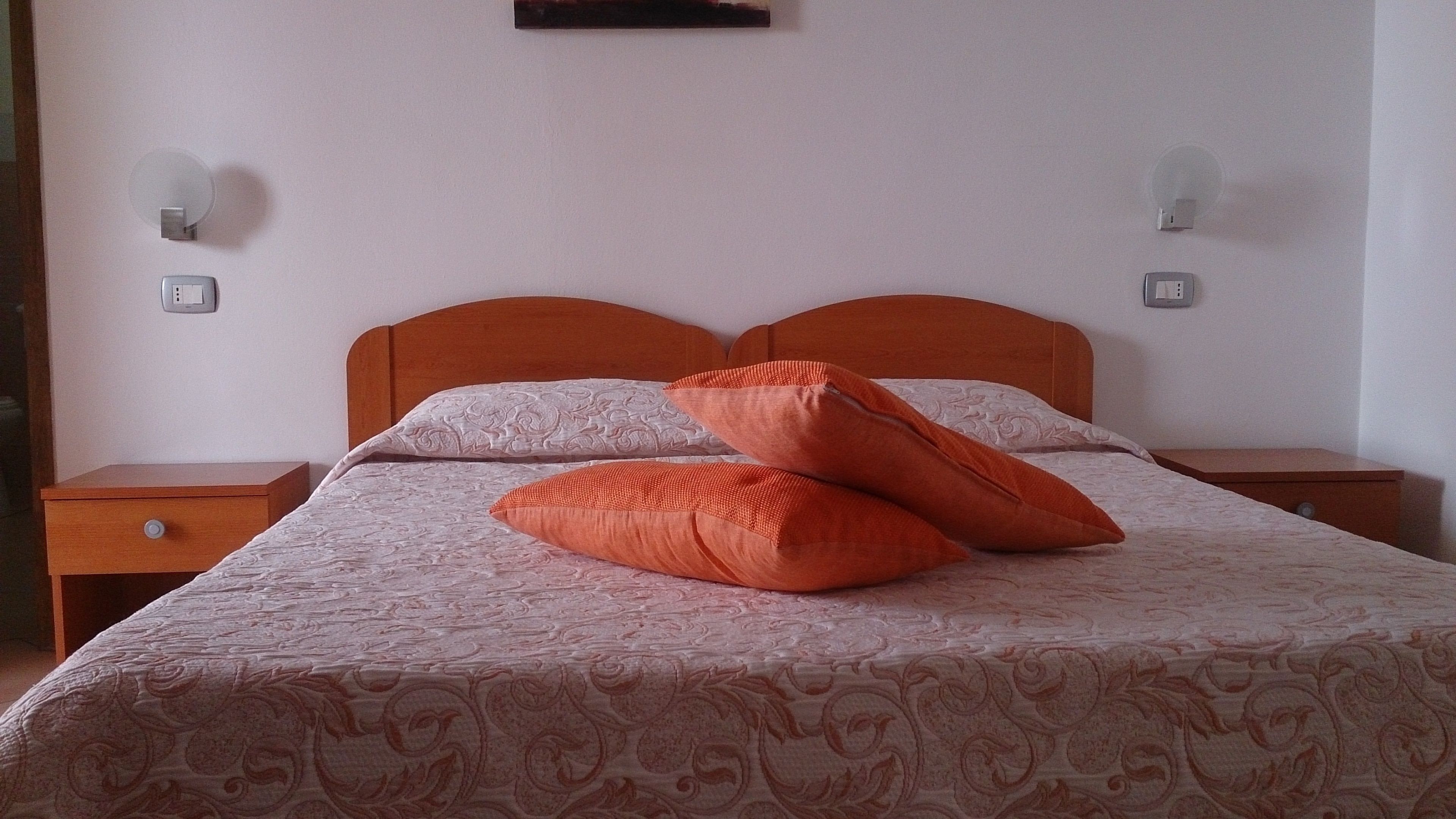 Un lit bien fait, avec une literie à motifs et deux oreillers orange, flanqué de deux tables de chevet en bois sur lesquelles sont posées des lampes murales.