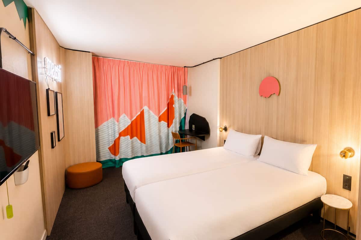 Habitación de hotel con una cama grande, un escritorio, una silla, un taburete pequeño, un perchero y un espejo.