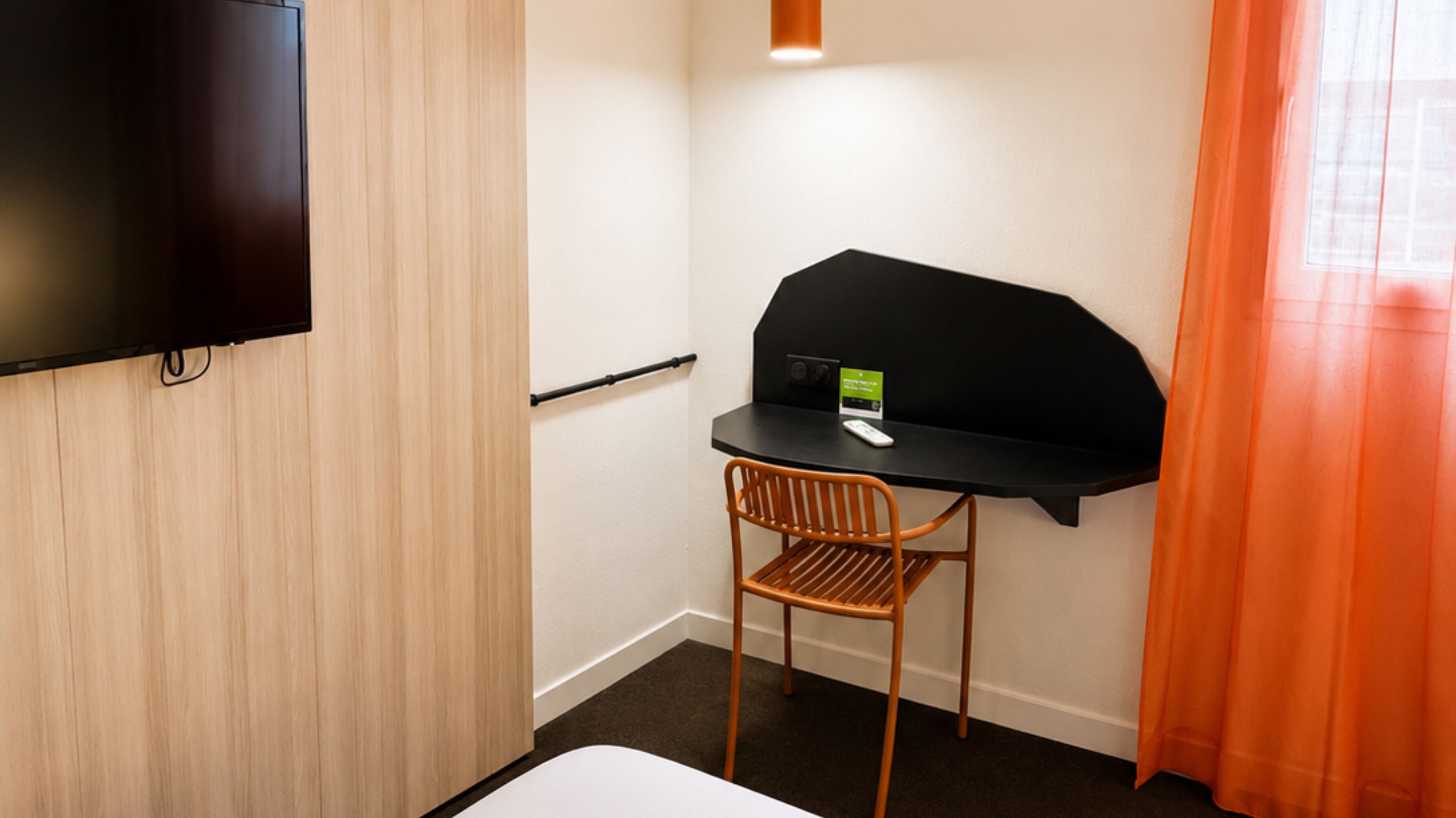 A hotel room with a bed, a desk with a chair, a television mounted on the wall, and a window with curtains.