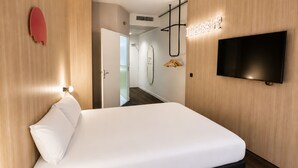 Una habitación de hotel con cama, televisor montado en la pared, puerta de madera y pasillo que lleva a un baño.