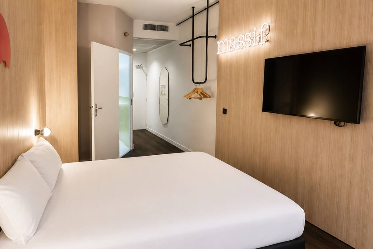 Una habitación de hotel con cama, televisor montado en la pared, puerta de madera y pasillo que lleva a un baño.